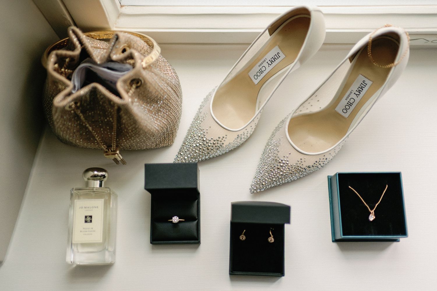Bridal flat lay featuring Jimmy Choo heels, Jo Malone perfume, diamond ring, gold necklace, and beaded clutch bag.