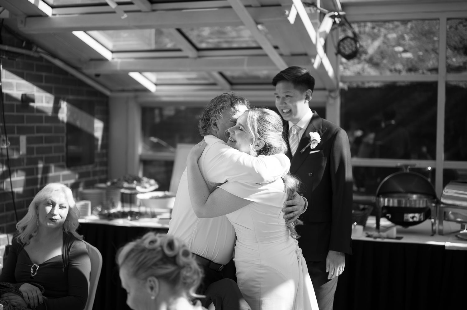 Black and white wedding photo of bride embracing guest while groom watches during indoor reception celebration.