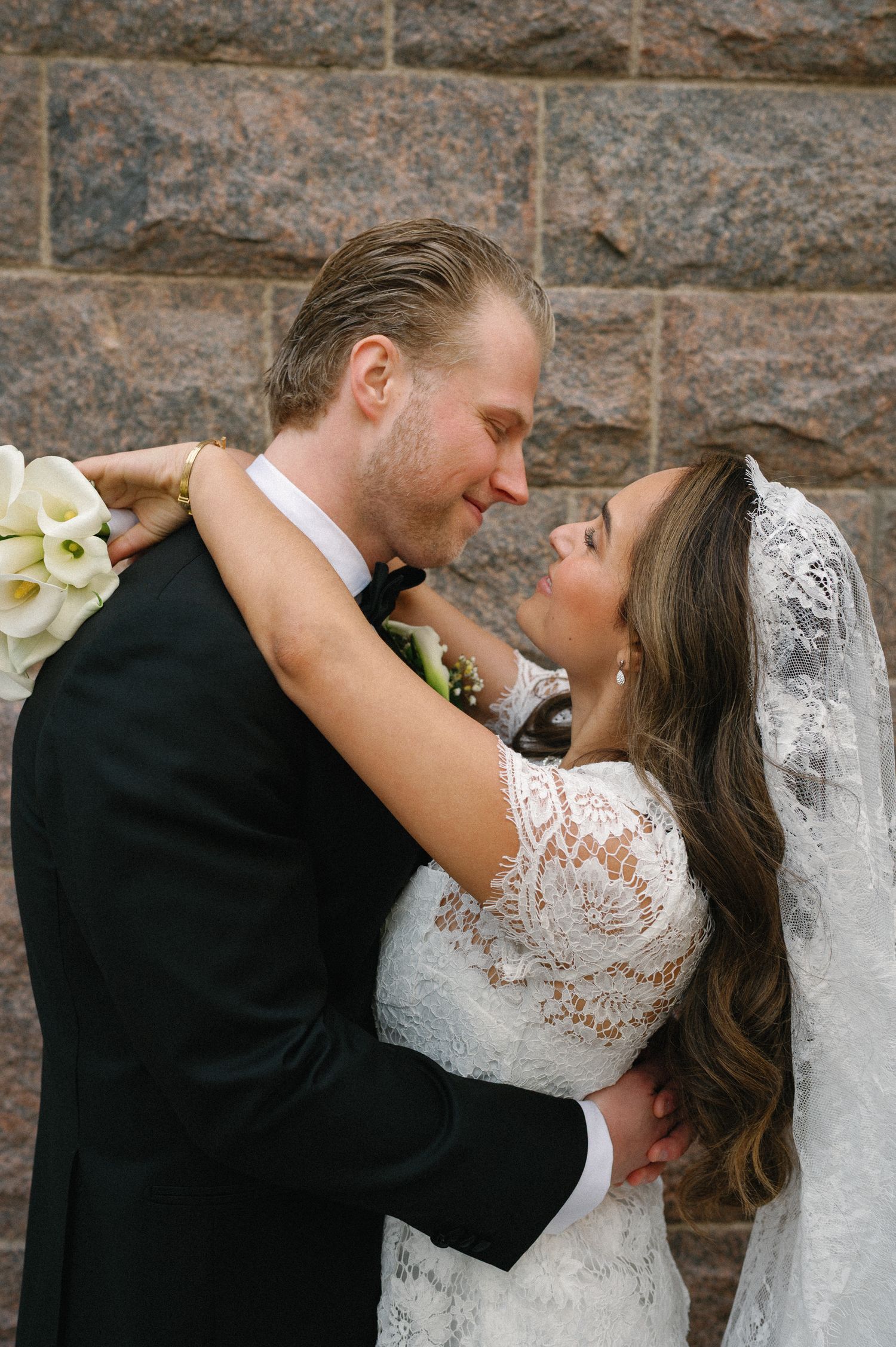 A bride in lace gown and veil embraces groom in black suit, sharing an intimate moment against a stone wall.