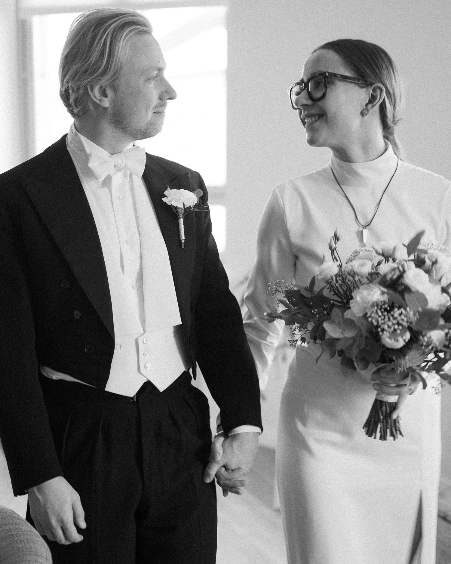 Black and white photo of a couple in wedding attire, groom in tuxedo and bride holding bouquet, smiling at each other.
