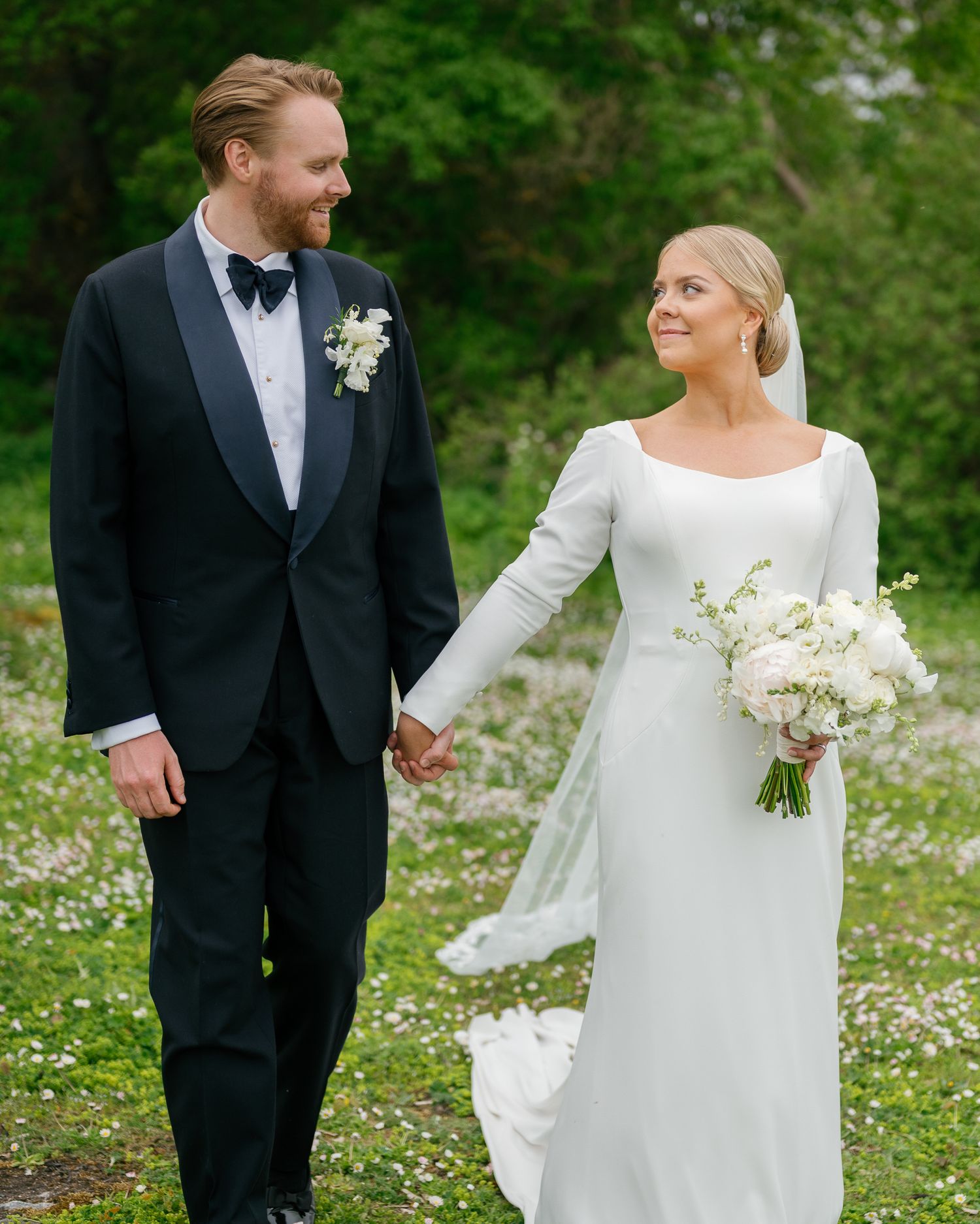 A bride and groom holding hands in a flower-filled meadow, she in a white gown with bouquet, he in a black tuxedo.