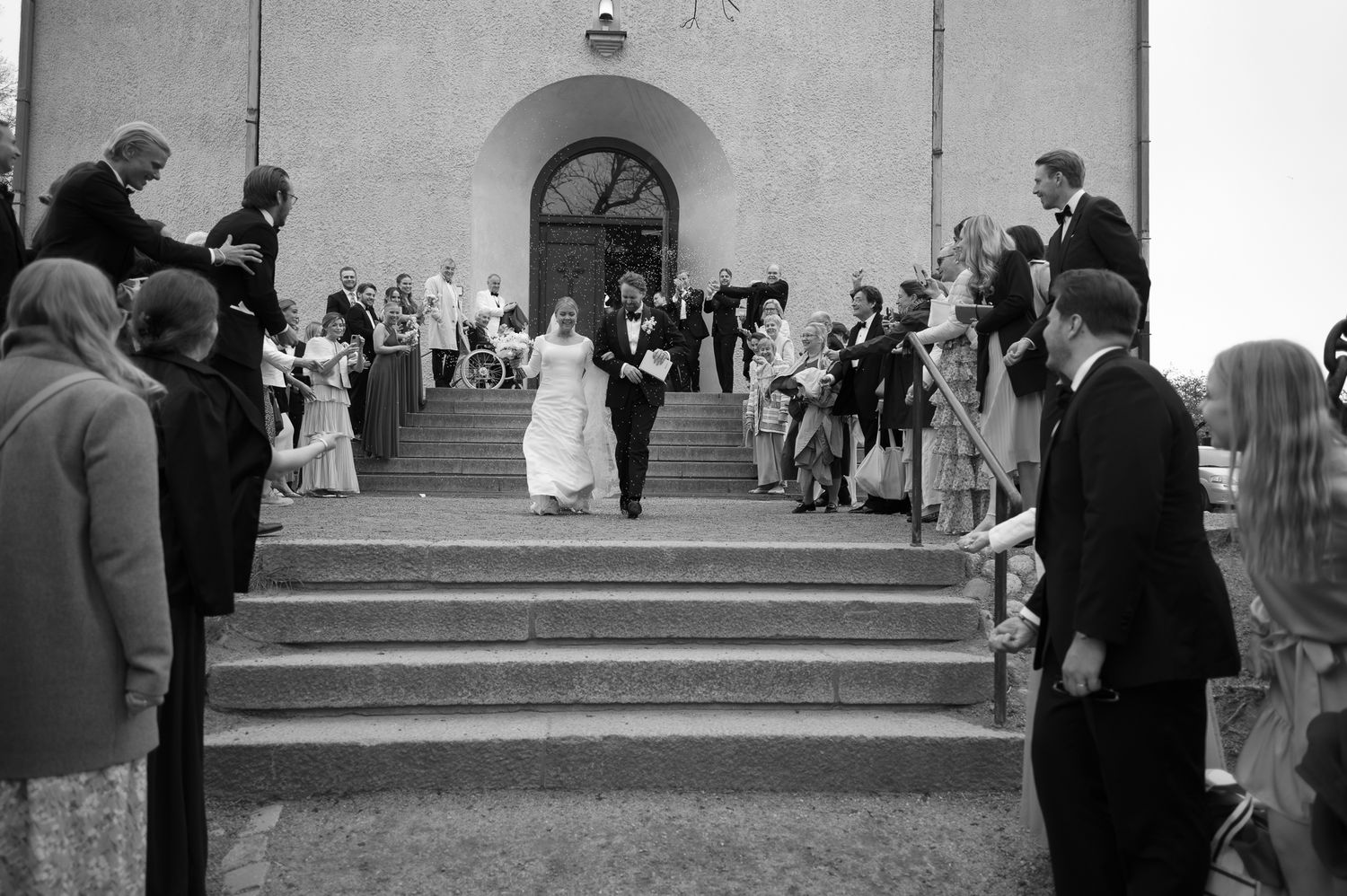 A black and white wedding photo showing a couple exiting a church while guests line the steps to celebrate.