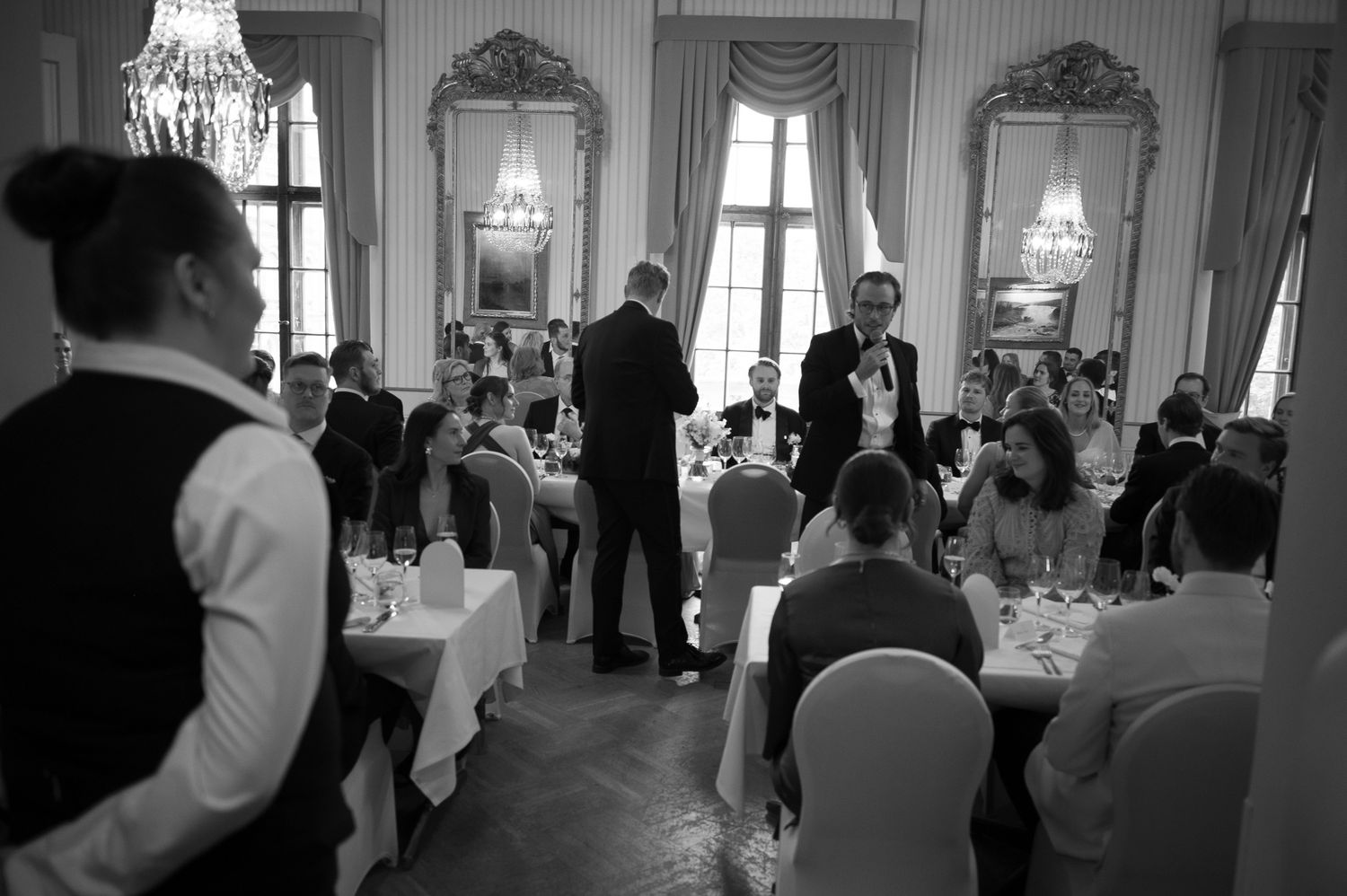 Black and white panorama of a formal event in an elegant ballroom with chandeliers and dining tables.