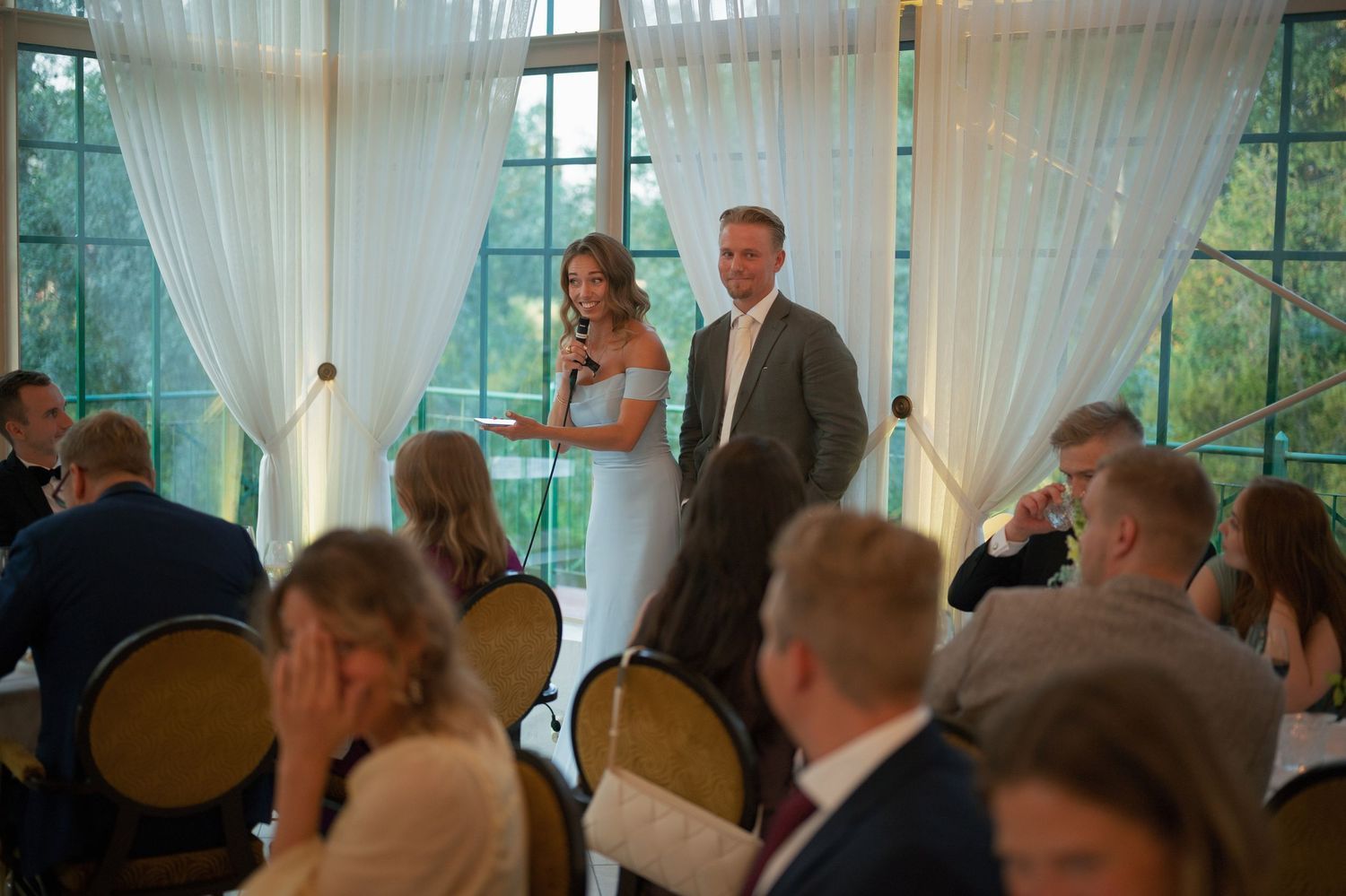 Wedding guests gathered in an elegant venue with white drapes and large windows.