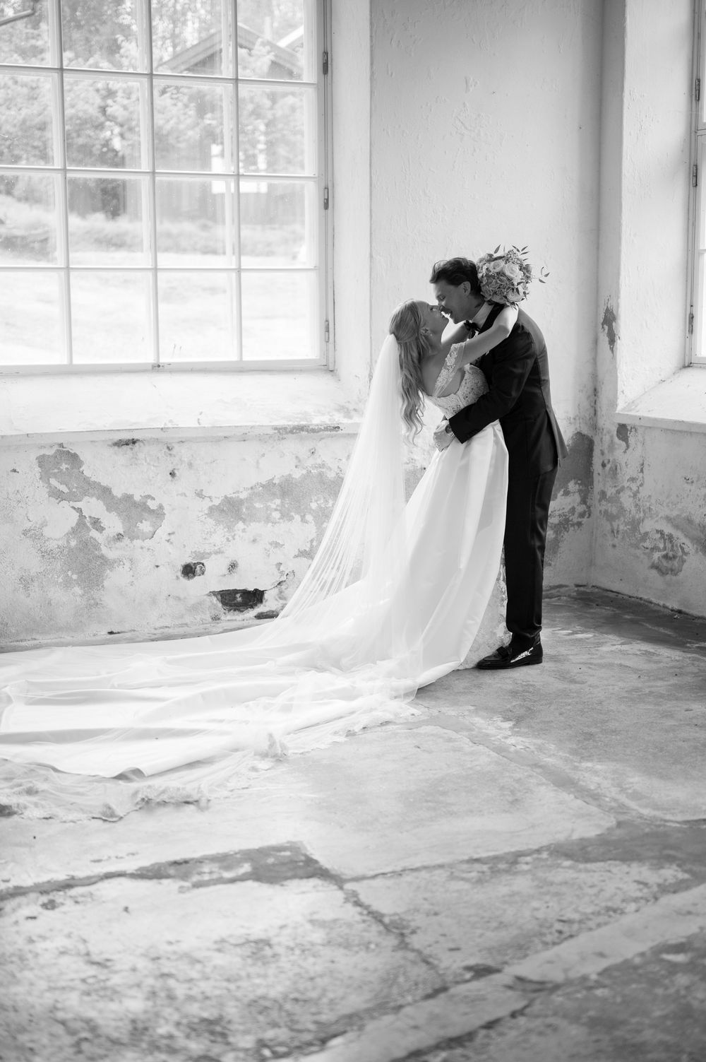 A full-length wedding photo showing the couple's romantic embrace in a rustic room with peeling walls.