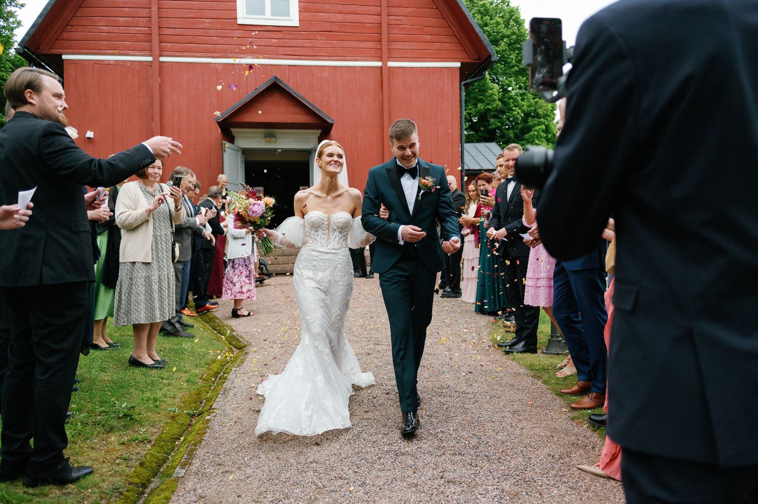 Newlyweds walk down path outside red church building surrounded by celebrating guests.