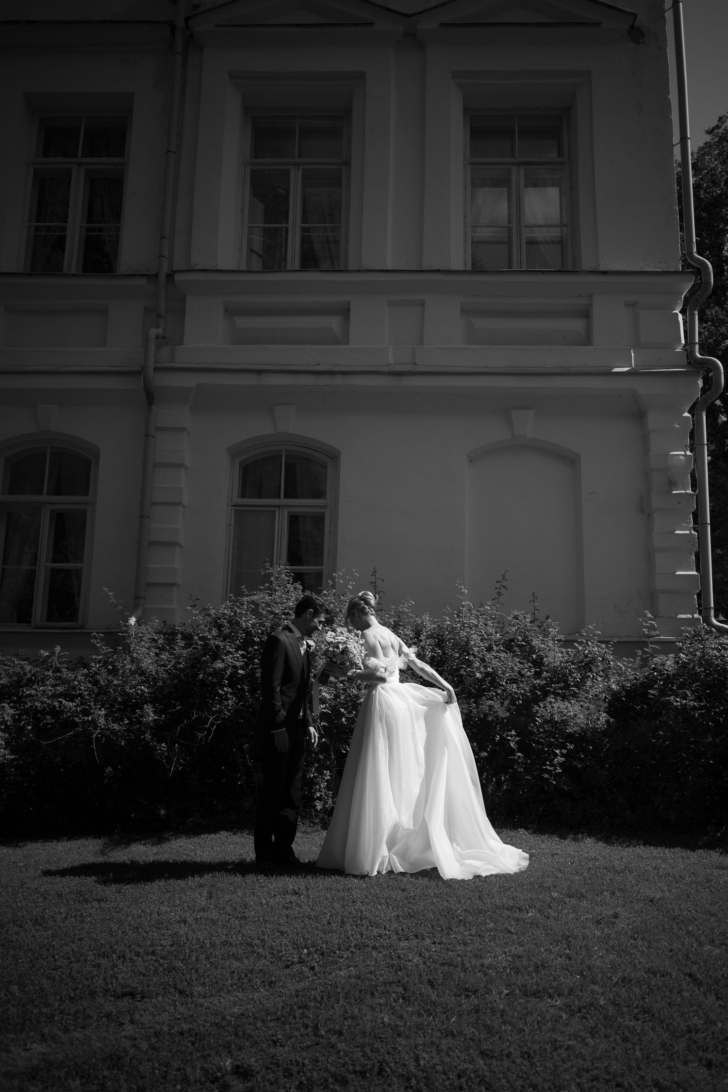 Black and white wedding photo of bride and groom standing together in front of elegant building surrounded by lush bushes.