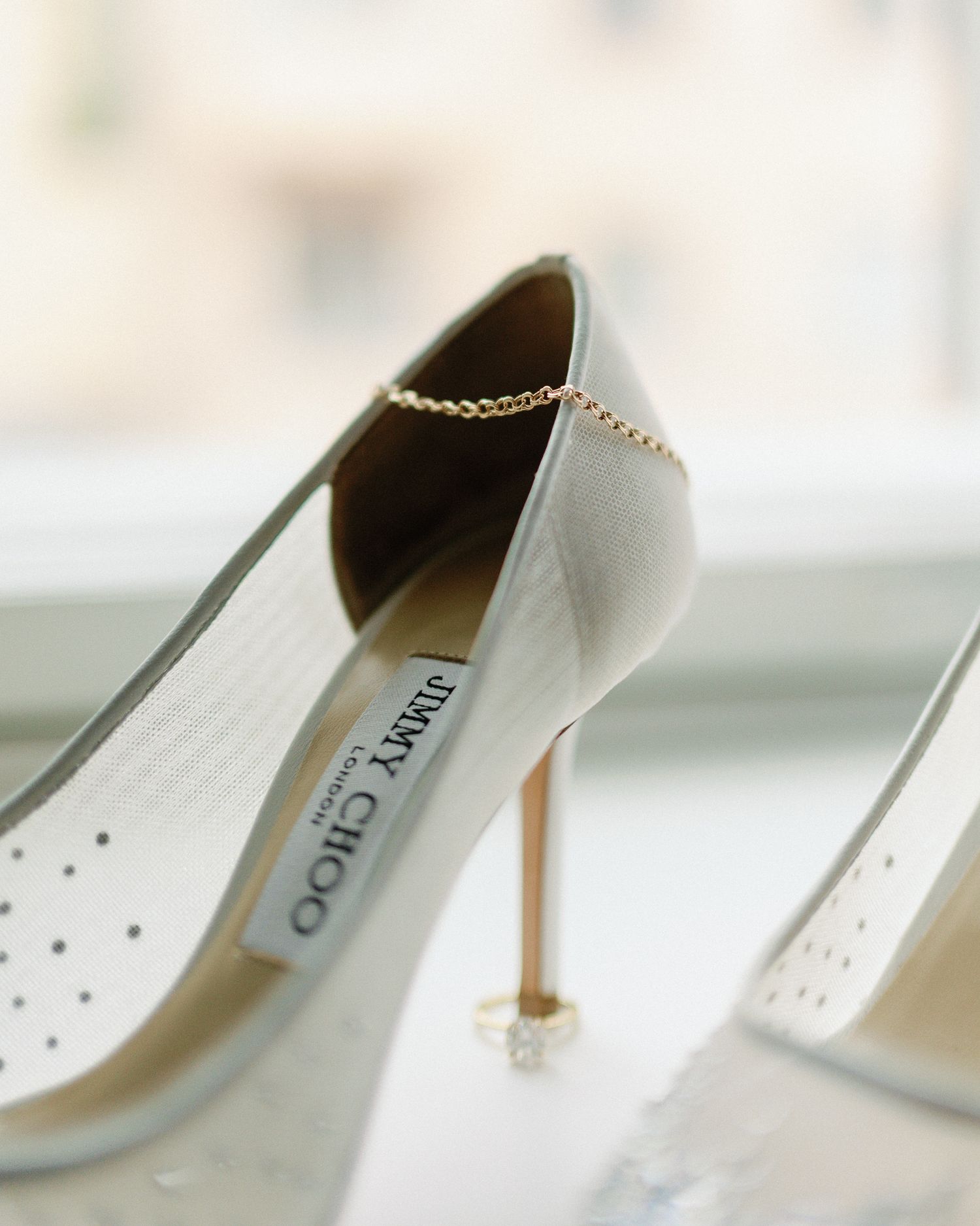 Elegant Jimmy Choo white satin heels with gold chain detail displayed on windowsill beside a diamond ring.
