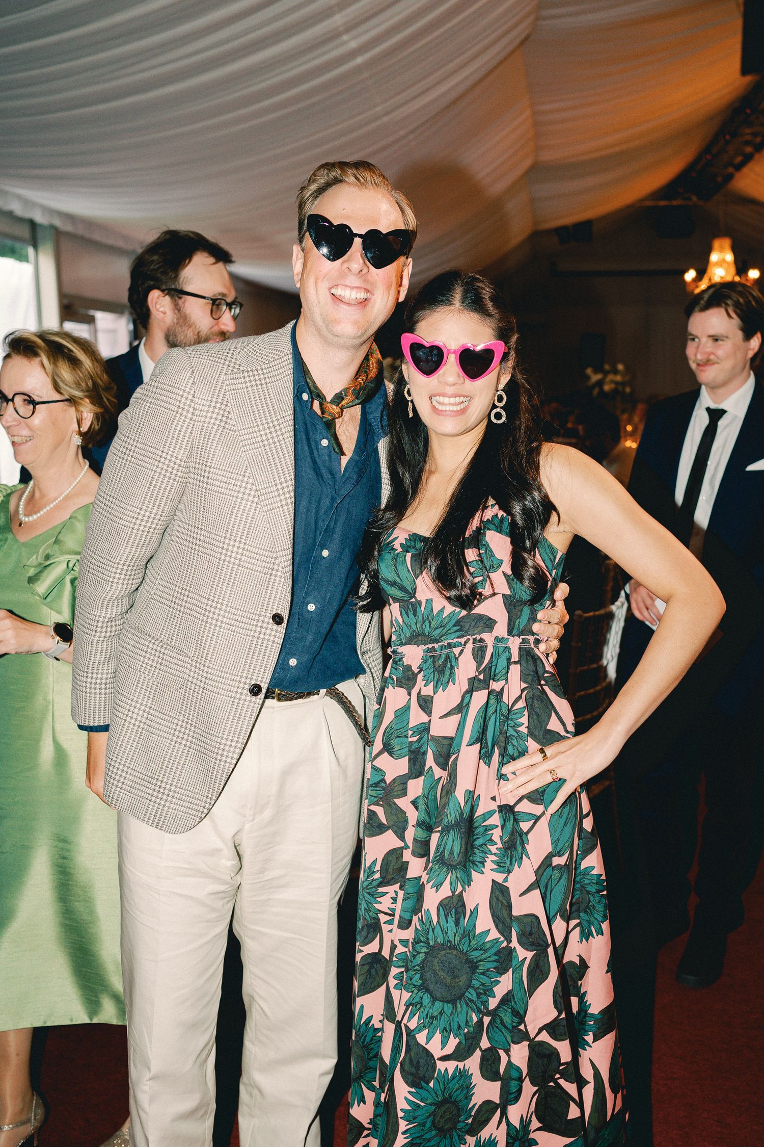 Couple wearing heart-shaped sunglasses posing together at a party, man in blazer, woman in floral dress.
