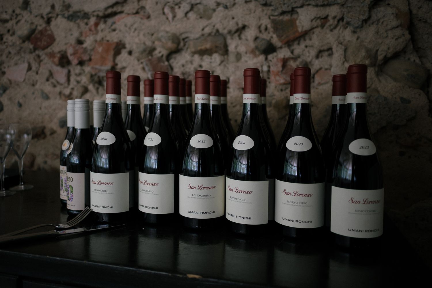 Rows of red wine bottles with white labels displayed on a dark shelf in a rustic stone cellar.