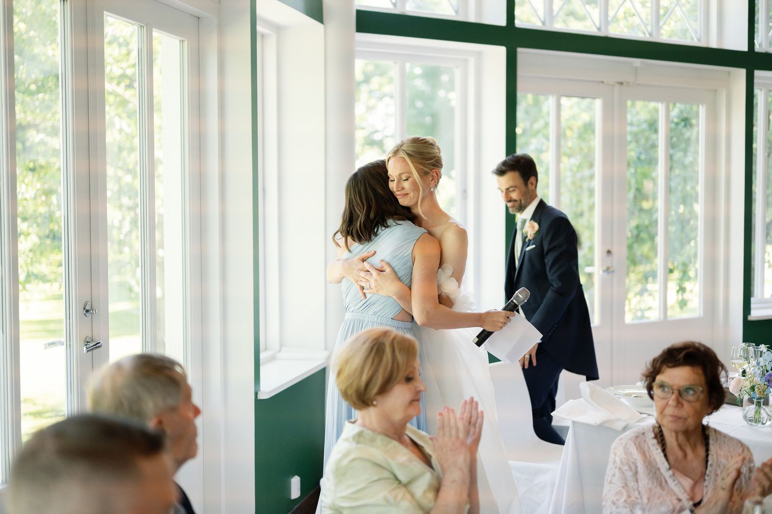 Two women share a warm hug at an elegant wedding reception while guests applaud and a groom stands nearby.