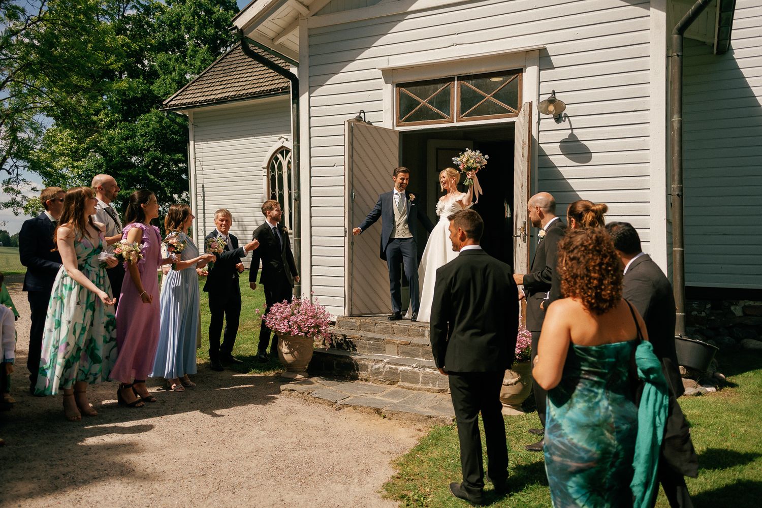 Wedding guests gather outside a white chapel as a couple exits their ceremony on a sunny day.