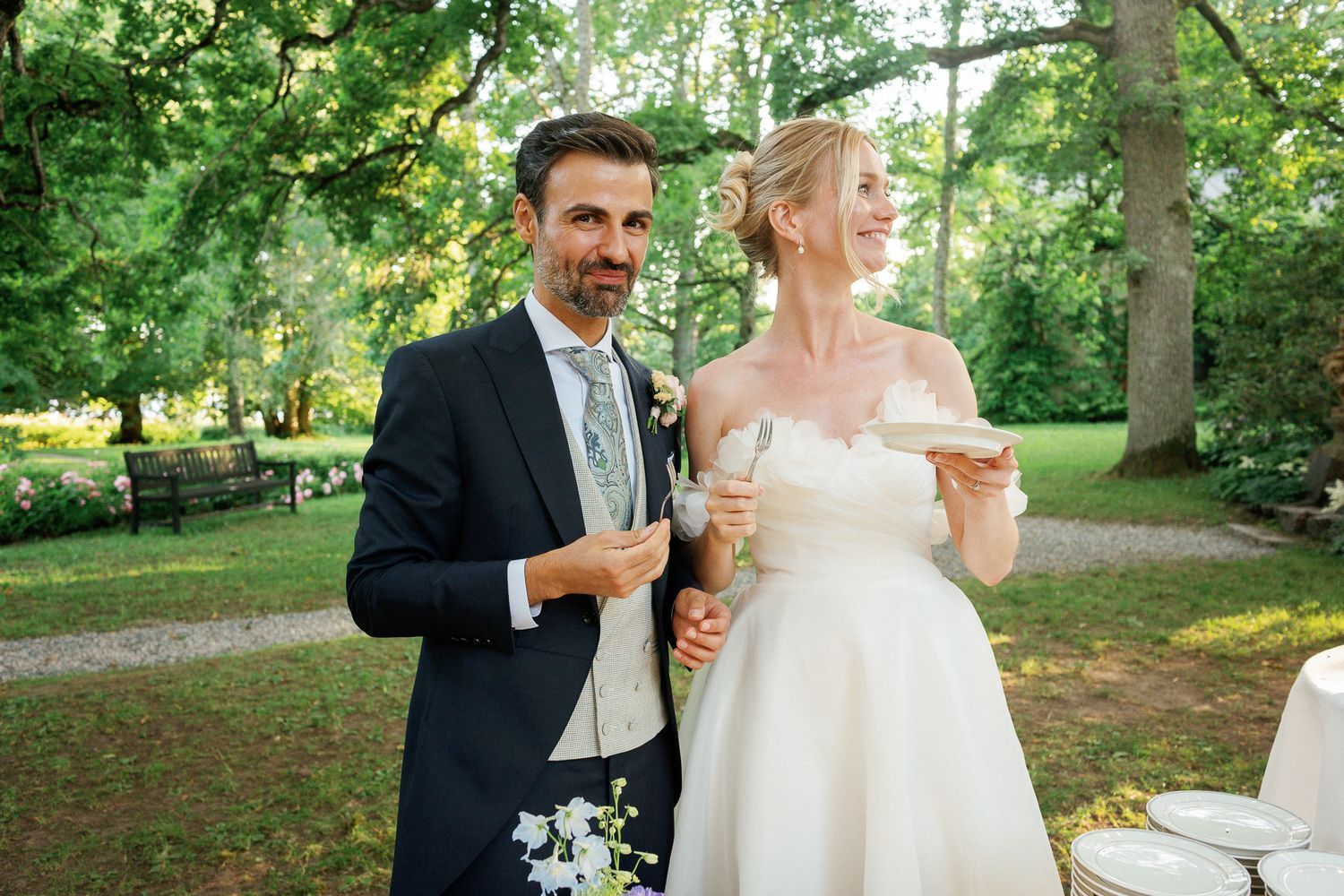 Bride in strapless white gown and groom in navy suit smile outdoors at elegant garden wedding ceremony.