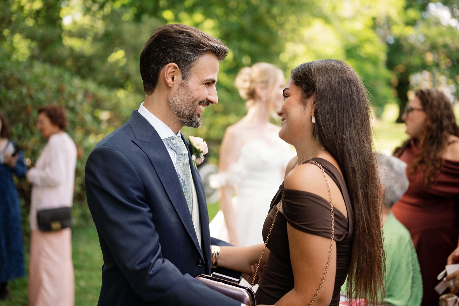 A couple smiles at each other during an outdoor wedding reception, man in navy suit, woman in brown dress.