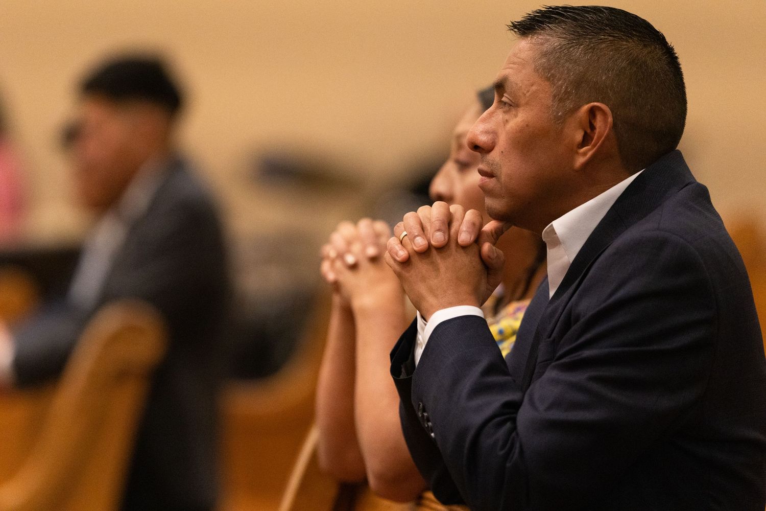 Man in suit praying with clasped hands in church pew during religious service.
