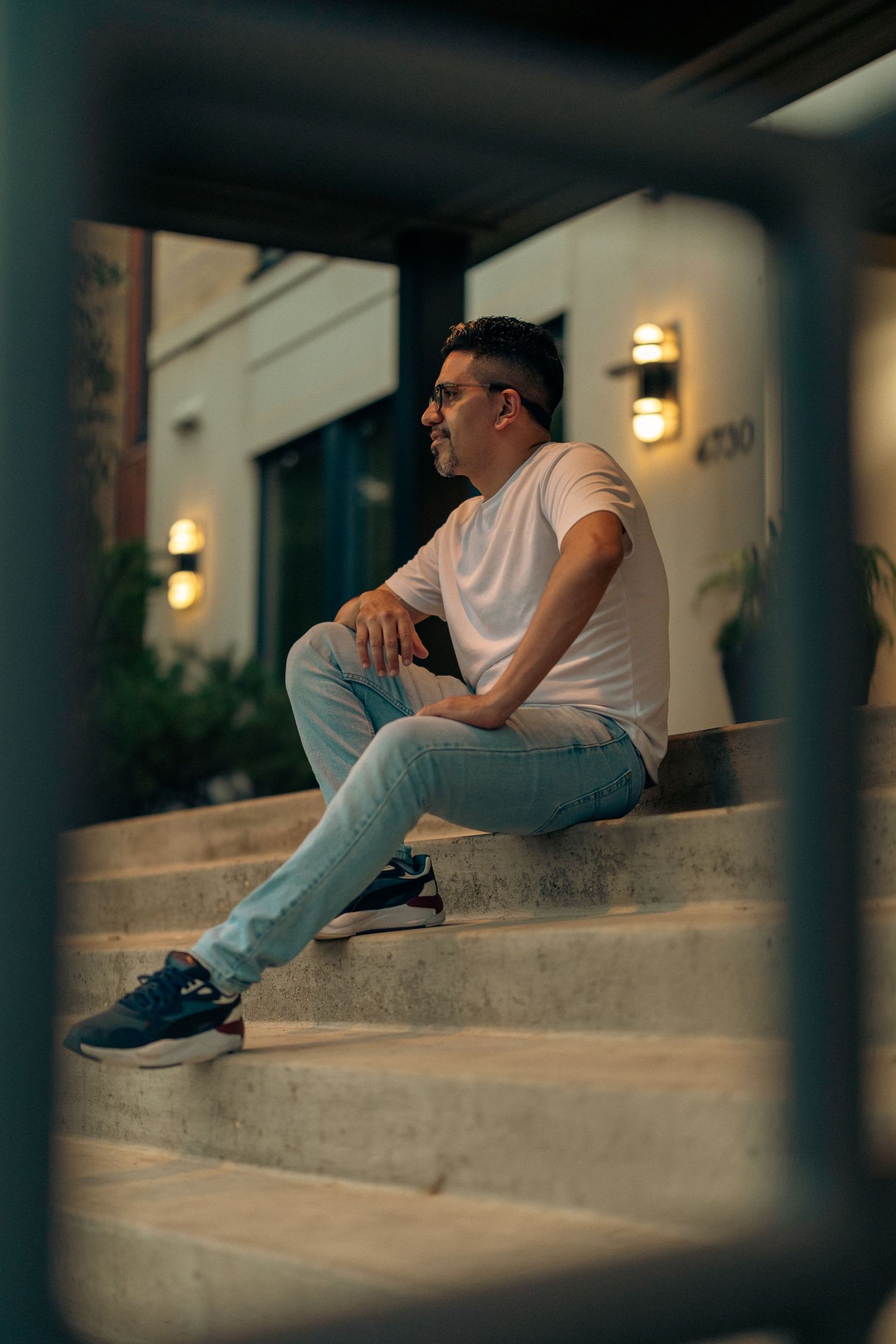 Man in white t-shirt and jeans sitting on outdoor steps at night near illuminated building entrance.