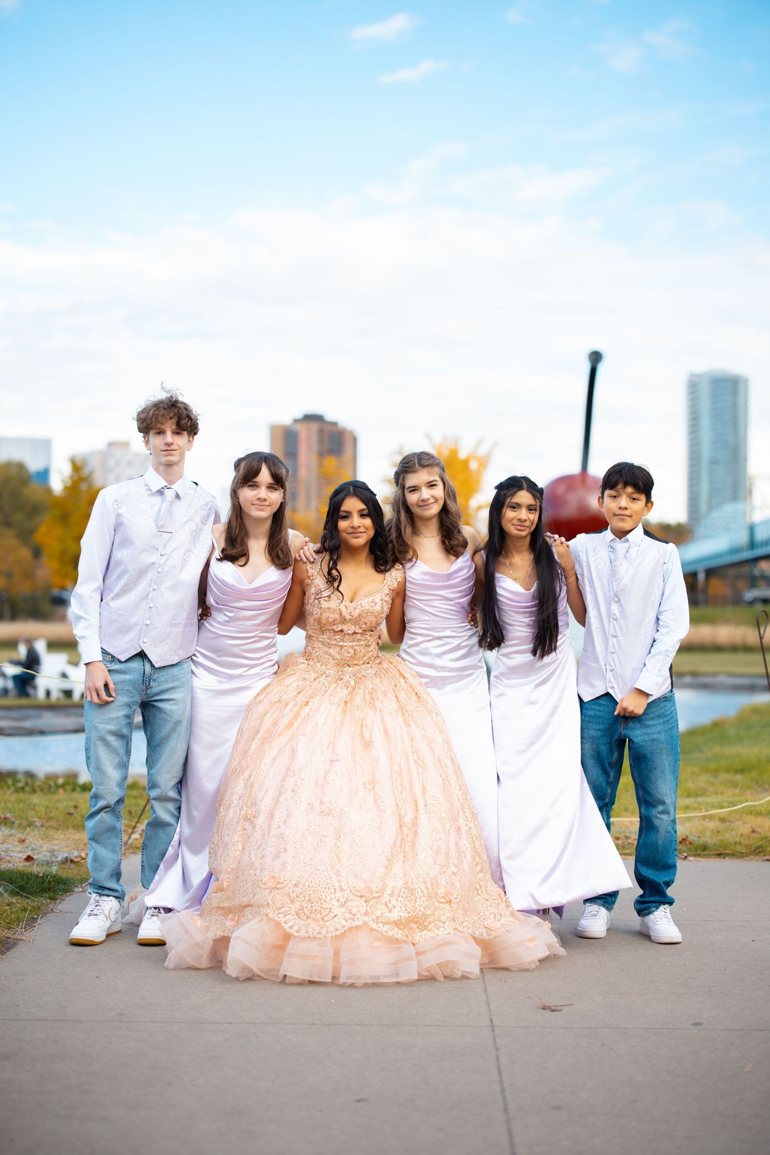Quinceañera girl in rose gold gown poses with four elegantly dressed court members near a city waterfront.