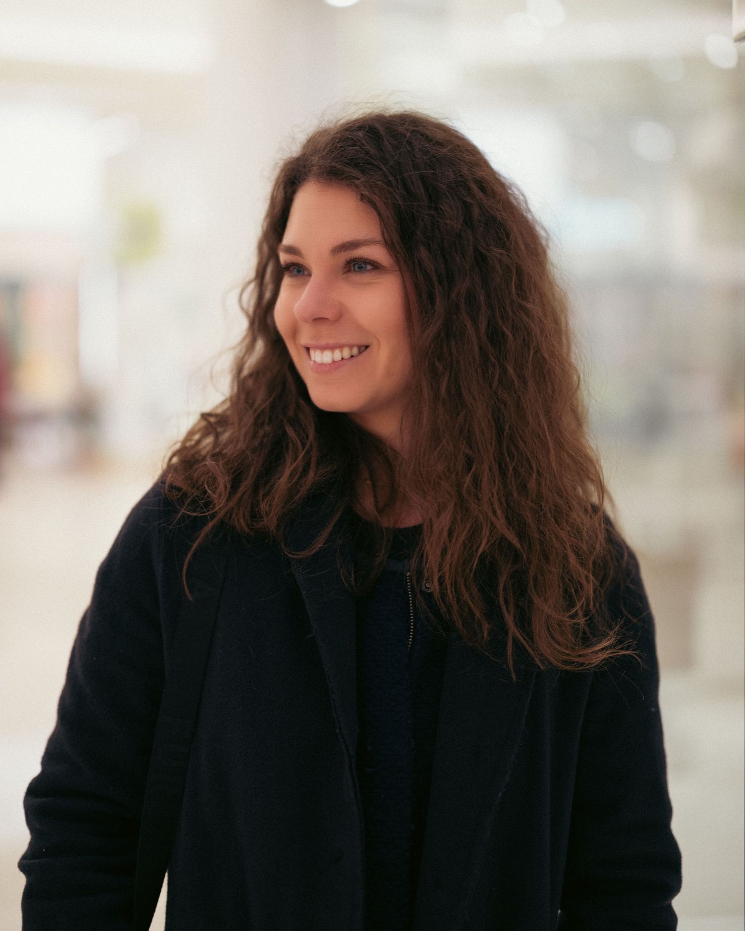 Smiling woman with curly brown hair wearing a black jacket in a bright indoor setting.
