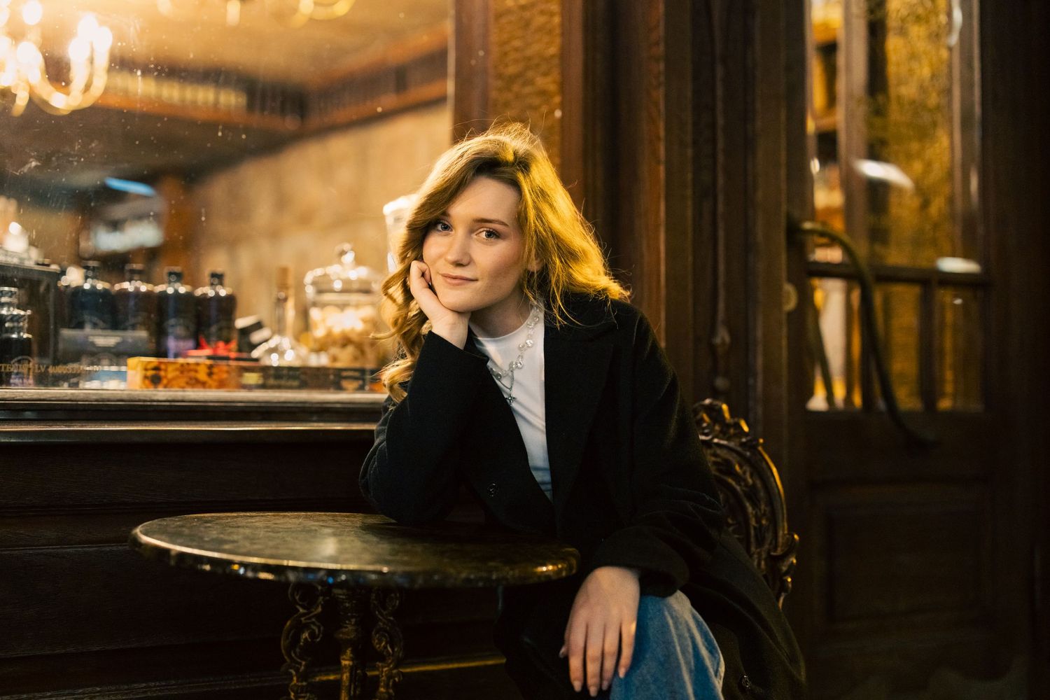 Young blonde woman sitting at a vintage cafe table, wearing a dark coat, in a warmly lit ornate bar setting.