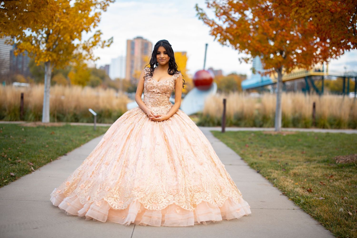 Young woman in elegant peach ball gown quinceañera dress poses on pathway surrounded by autumn foliage.