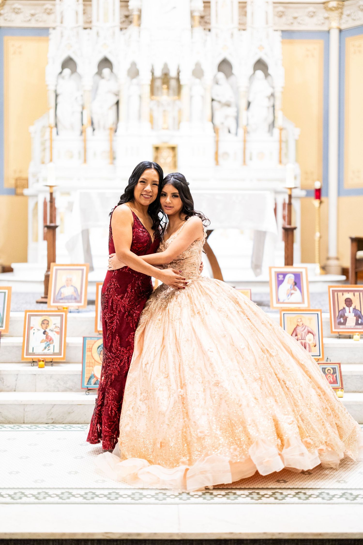Mother in red dress embraces daughter in peach quinceañera gown before white church altar backdrop.