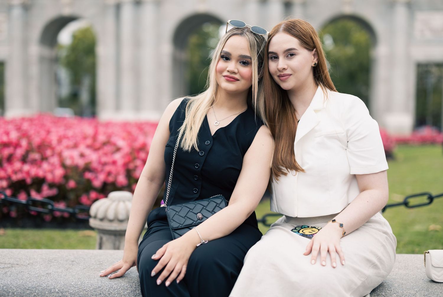 Two young women sitting together outdoors near pink flowers, one blonde in black dress, one brunette in white.