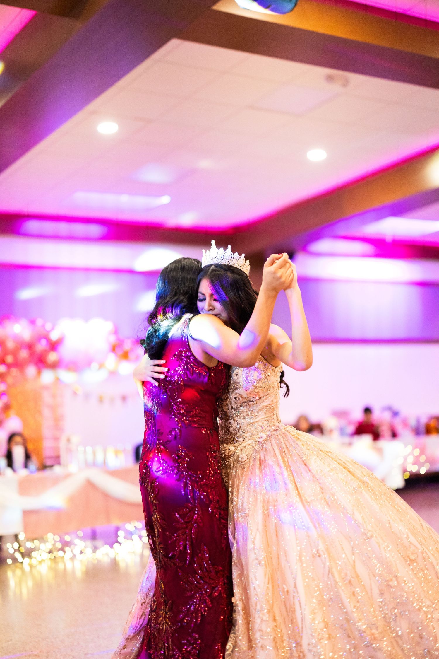 Two women in formal gowns share an emotional dance at a quinceañera celebration in a decorated ballroom.