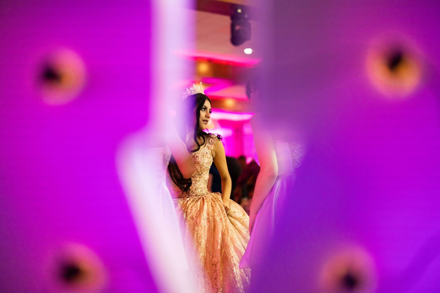 Quinceañera girl in peach gown and tiara photographed through decorative elements with purple lighting backdrop.