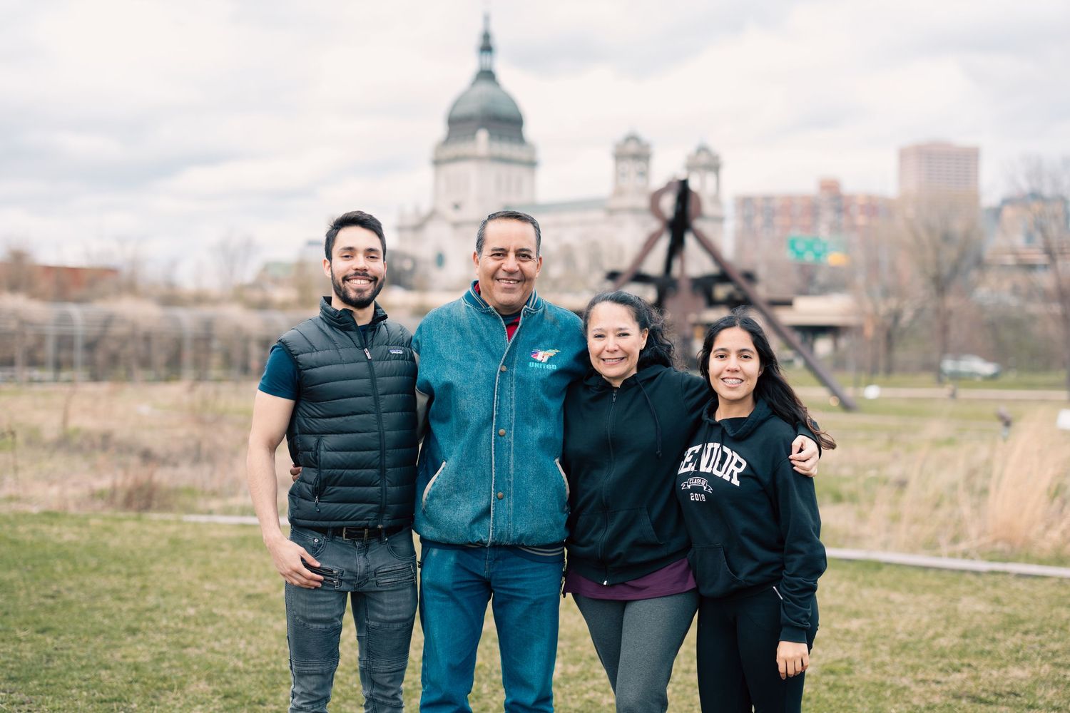 Family of four smiling together outdoors with a domed capitol building and sculpture in the background.