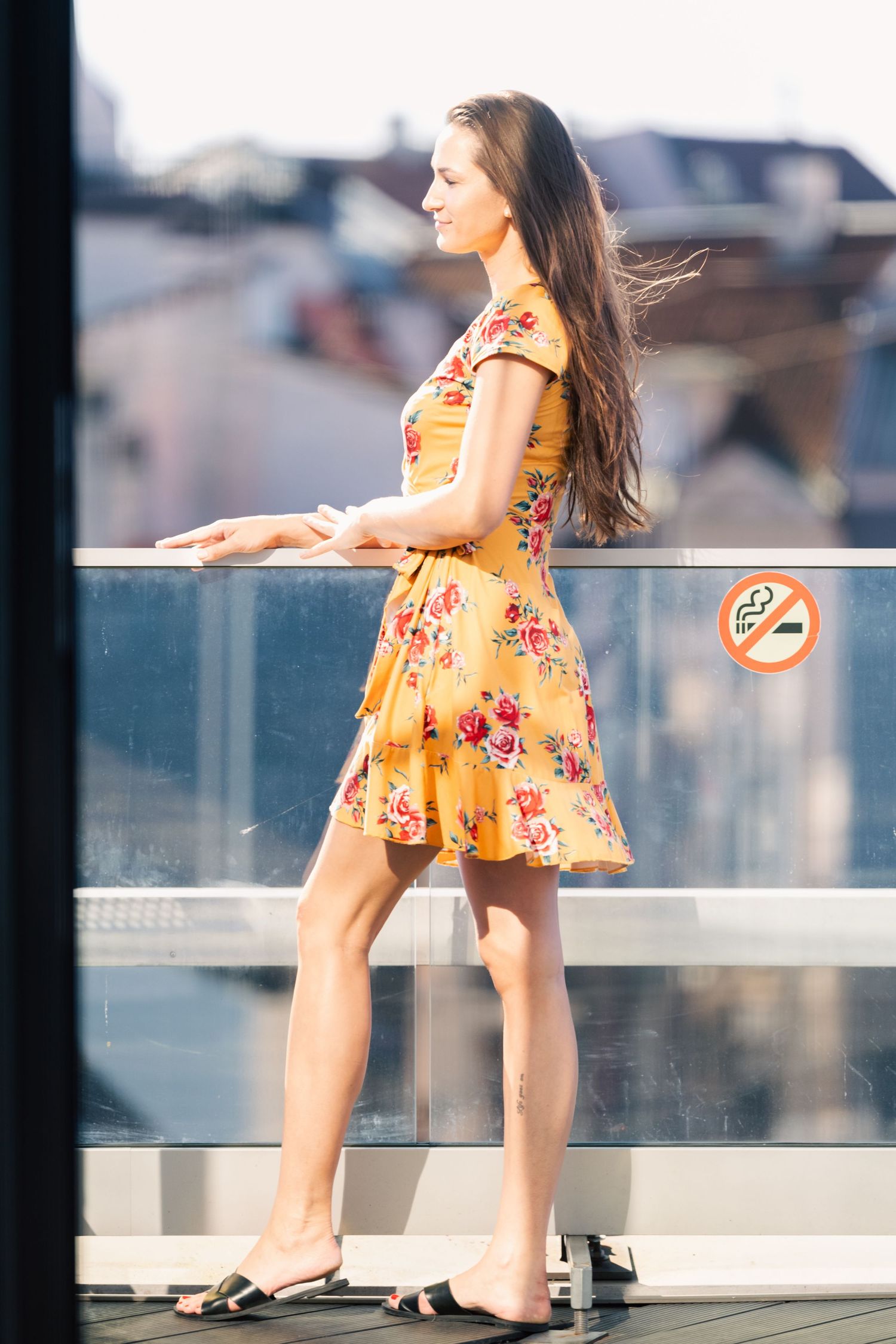 Young woman in yellow floral dress standing on rooftop terrace with city skyline in background.