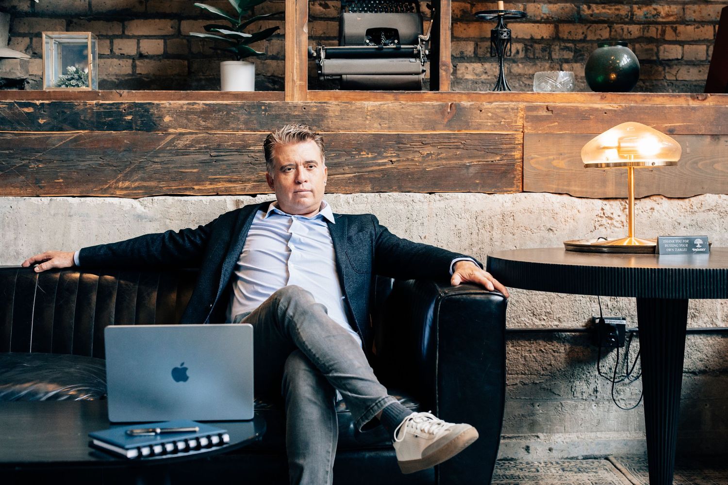 Confident businessman relaxing on leather sofa with MacBook laptop in modern industrial office space.