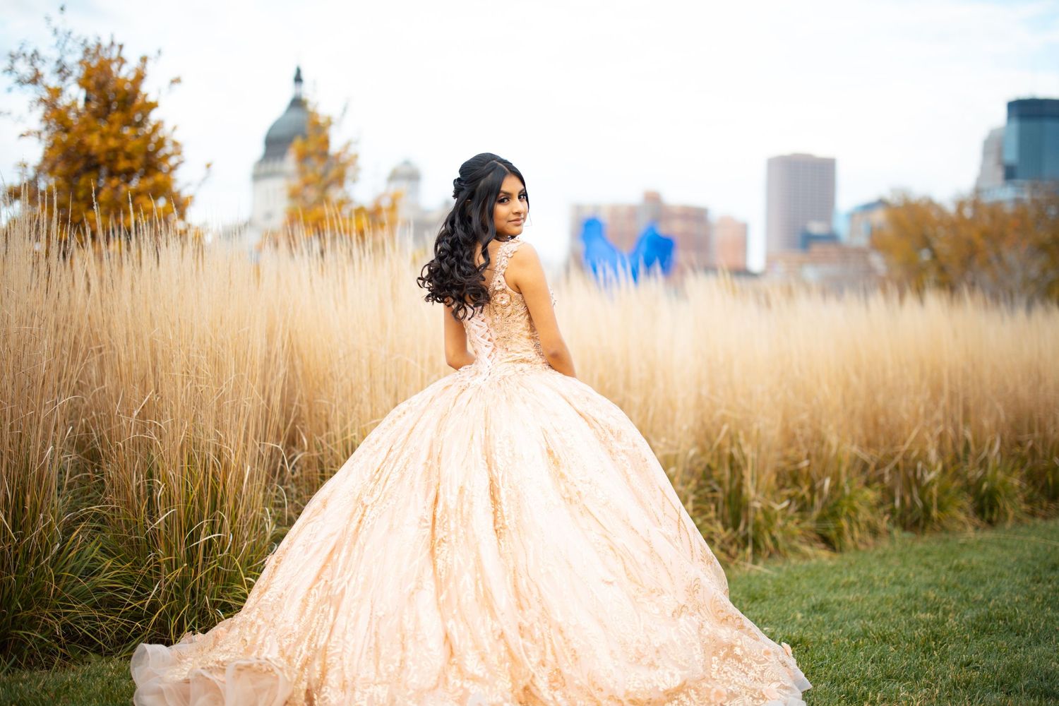 Young woman in elegant gold quinceañera ball gown poses in field with city skyline backdrop.