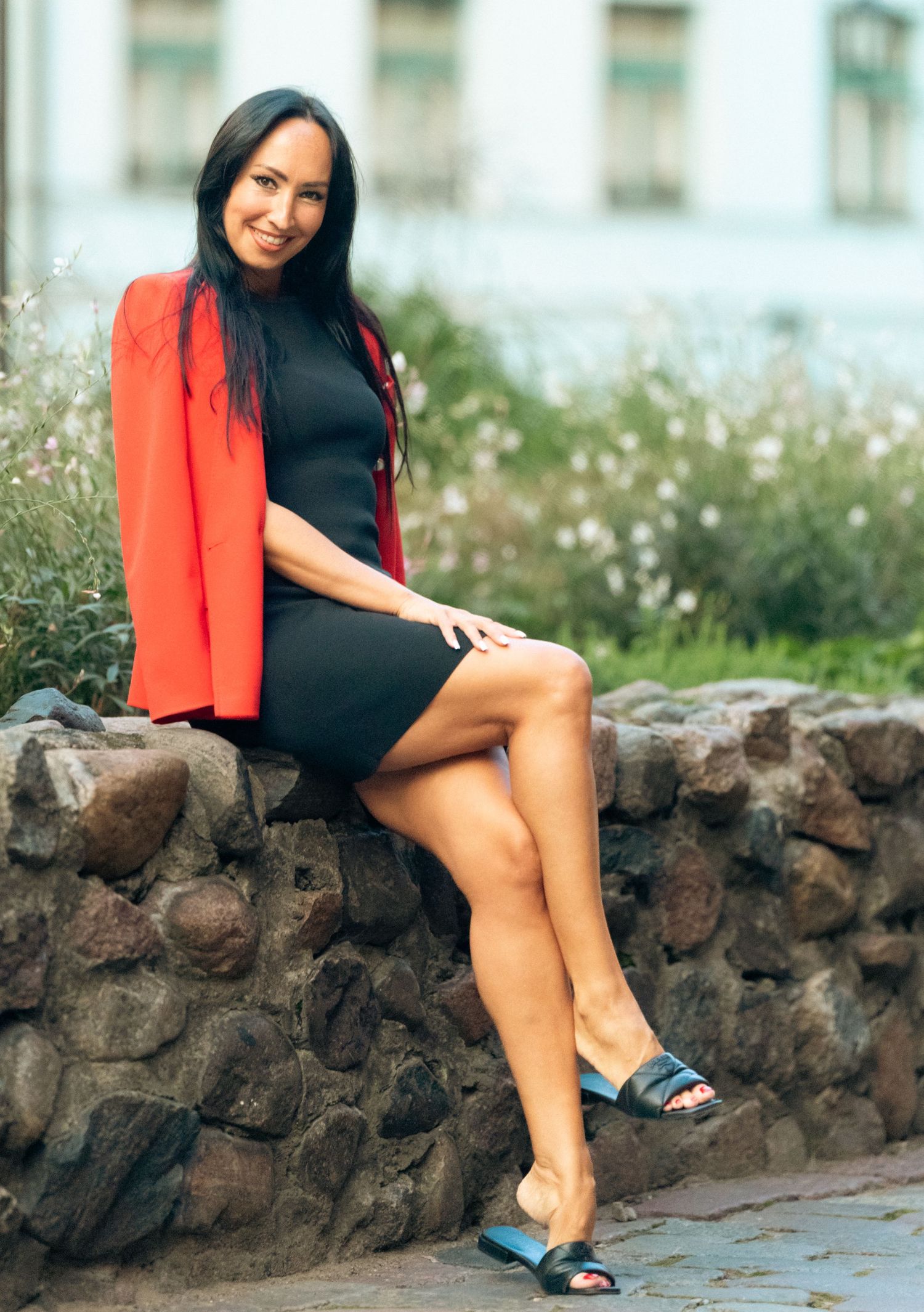 Brunette woman in black dress and red blazer sitting on stone wall outdoors smiling at camera.