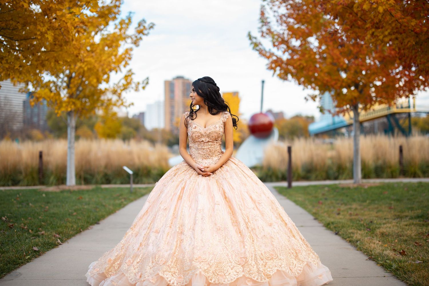 Young woman in a golden ball gown quinceañera dress poses on a park path surrounded by colorful autumn foliage.