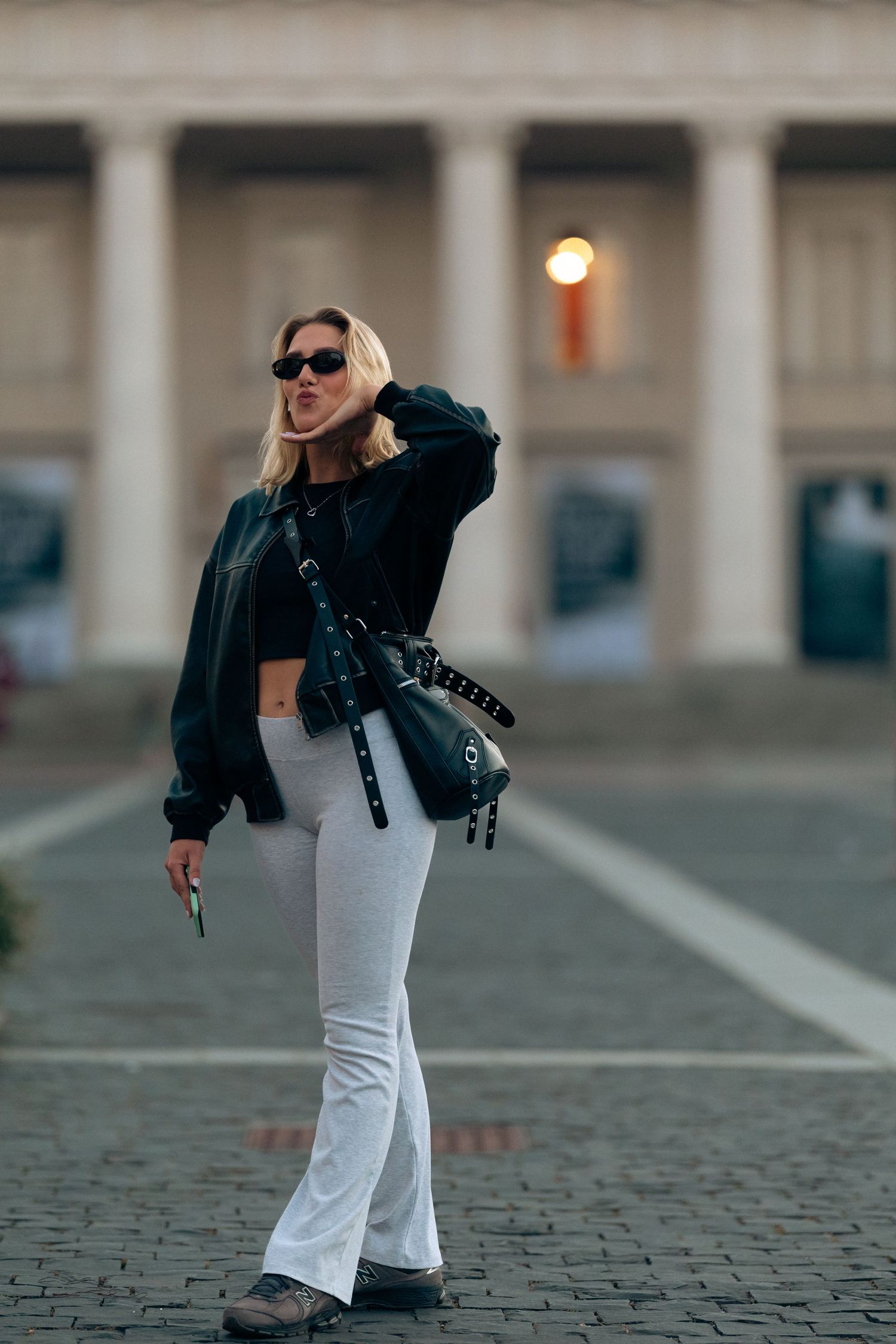 Stylish blonde woman in black jacket and white flared pants poses in sunglasses at city square.