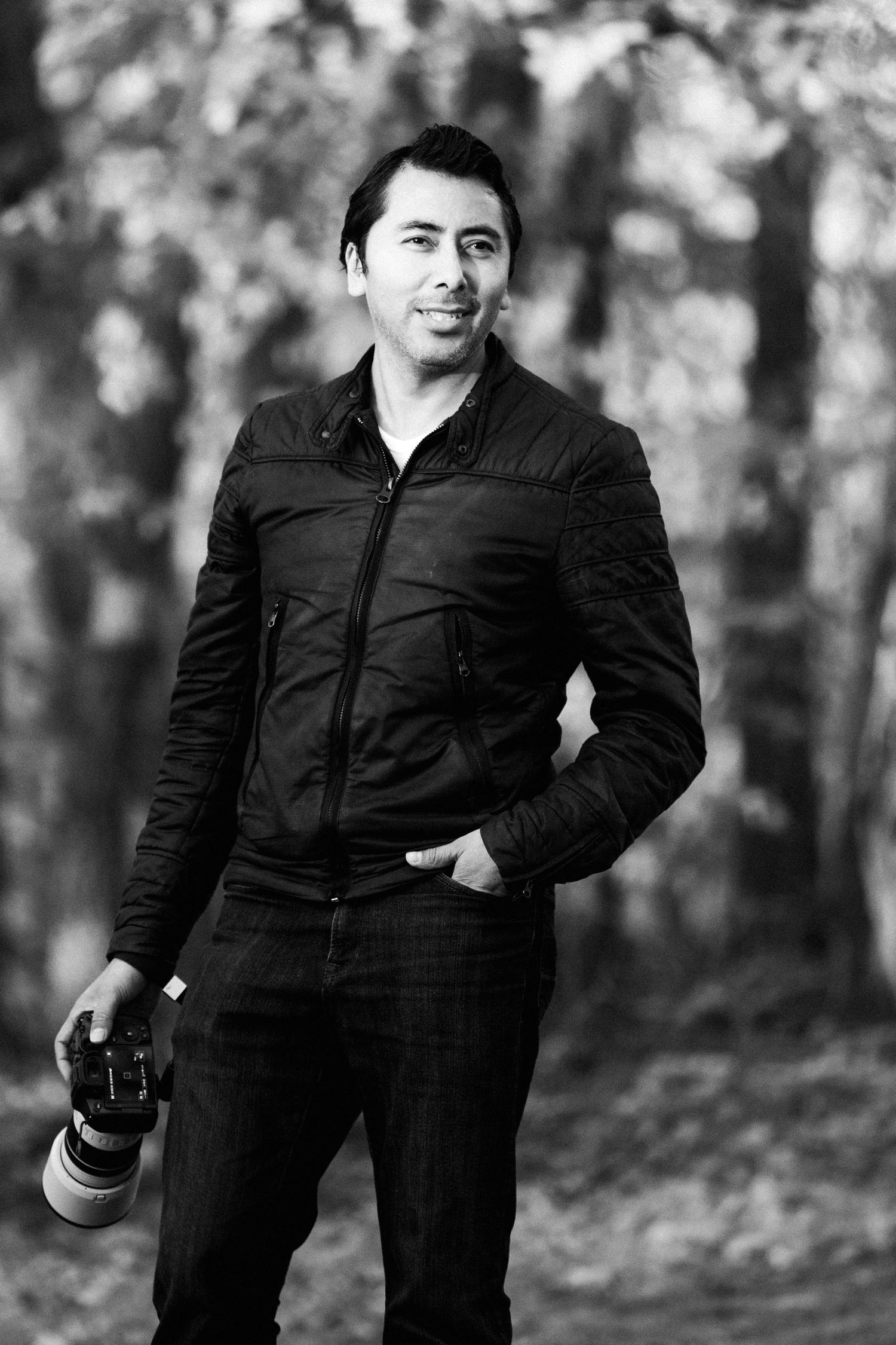 Black and white portrait of a man in a dark jacket holding a camera with a telephoto lens outdoors.