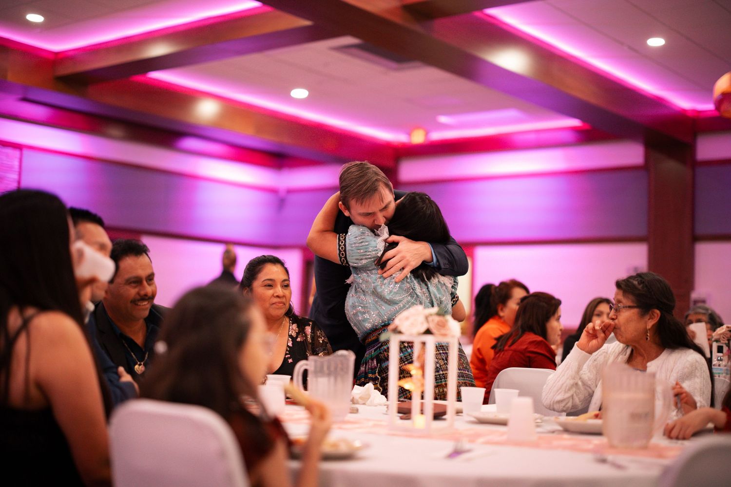 Emotional family moment as guests embrace at a quinceañera reception dinner in a pink-lit ballroom.