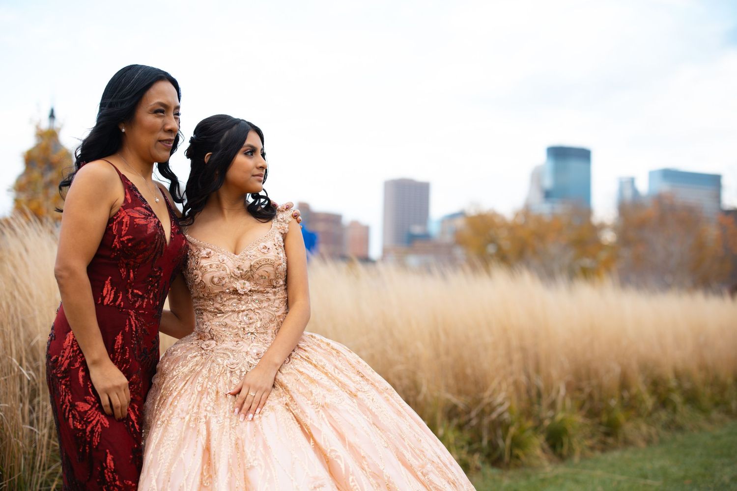 Two young women in formal gowns, one red sequined and one rose gold ball gown, pose before tall golden grasses.