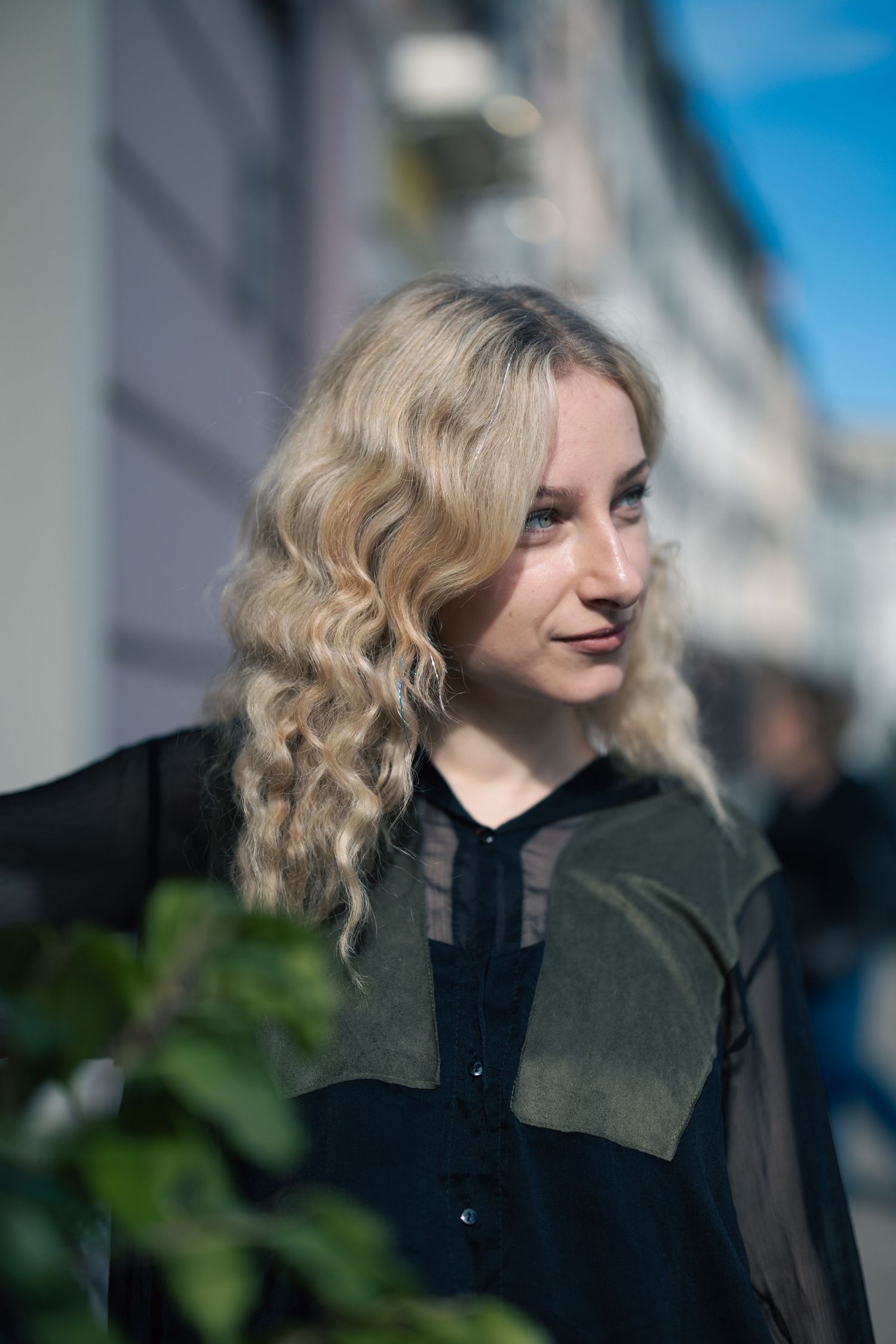 Blonde woman with curly hair wearing a black and olive shirt poses outdoors on a sunny urban street.