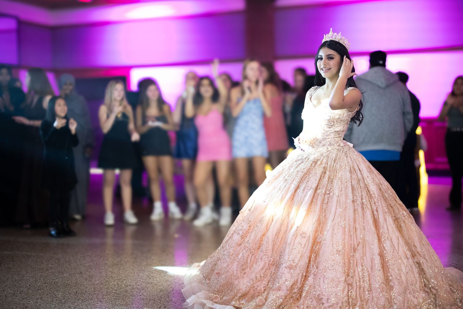Quinceañera girl wearing tiara and peach ball gown stands on dance floor surrounded by guests.