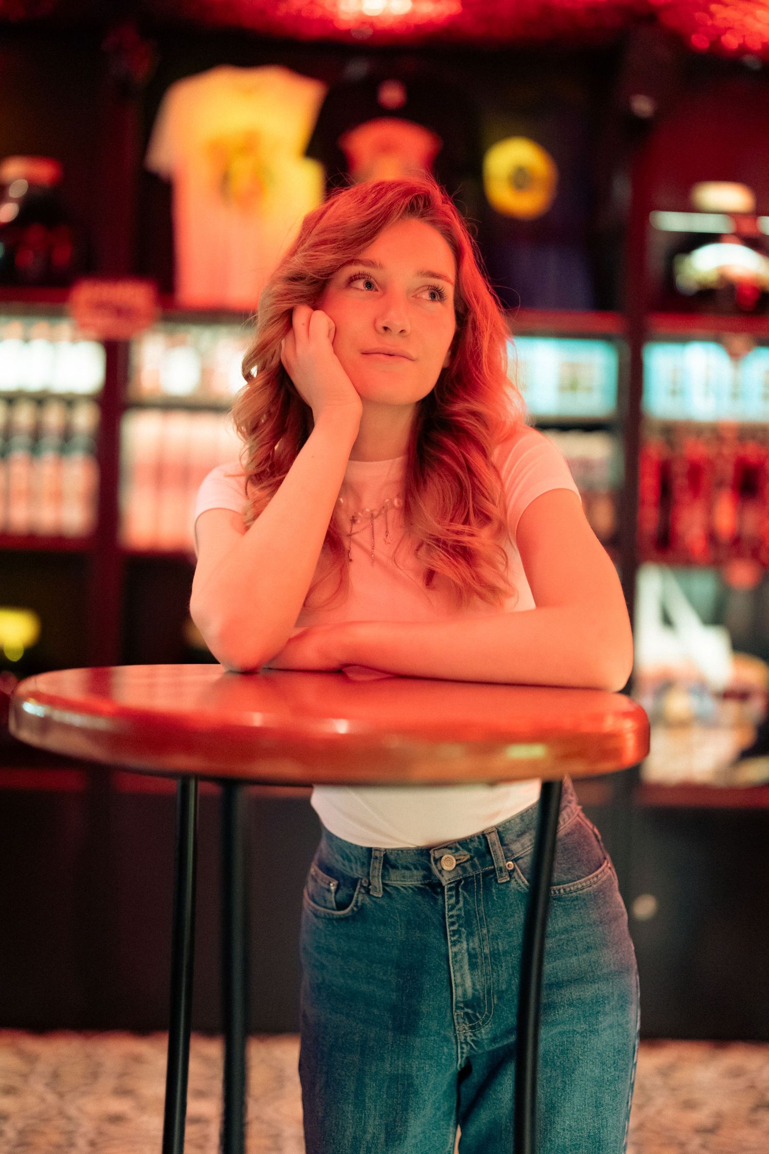 Young redhead woman rests chin on hands leaning on round table in red-lit bar interior.