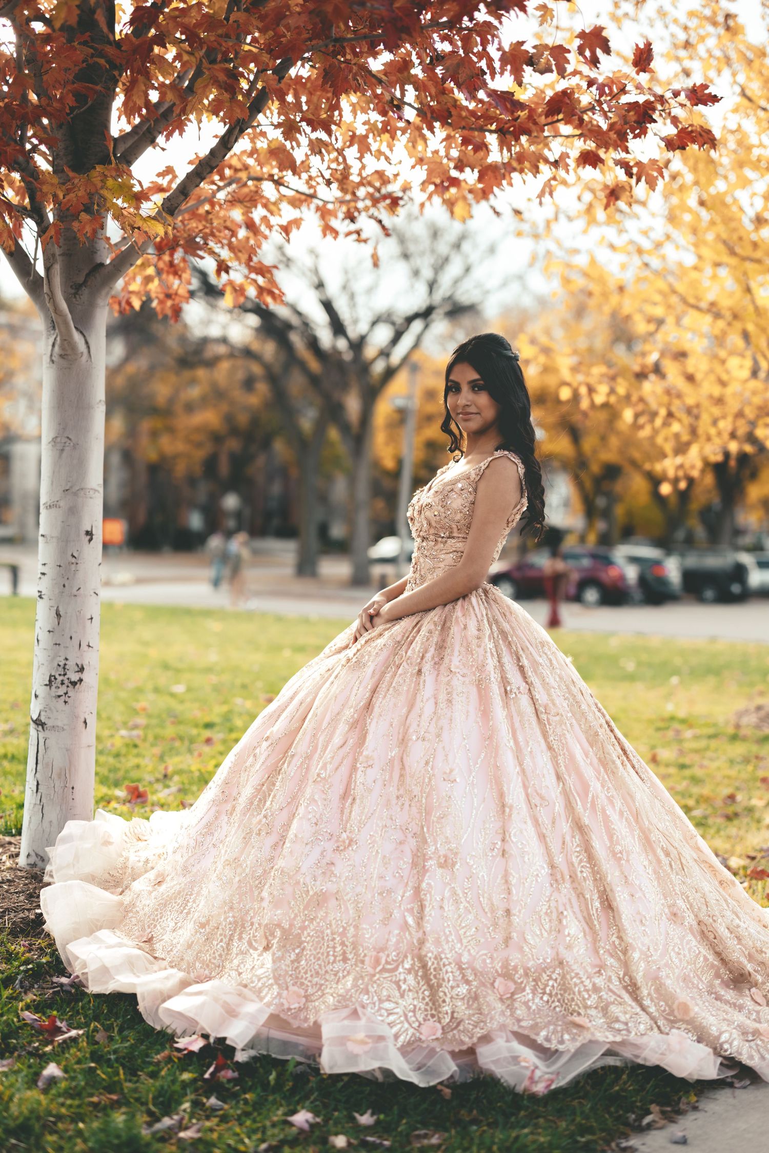 Smiling quinceañera girl in elaborate rose gold ball gown poses beside birch tree with autumn foliage backdrop.