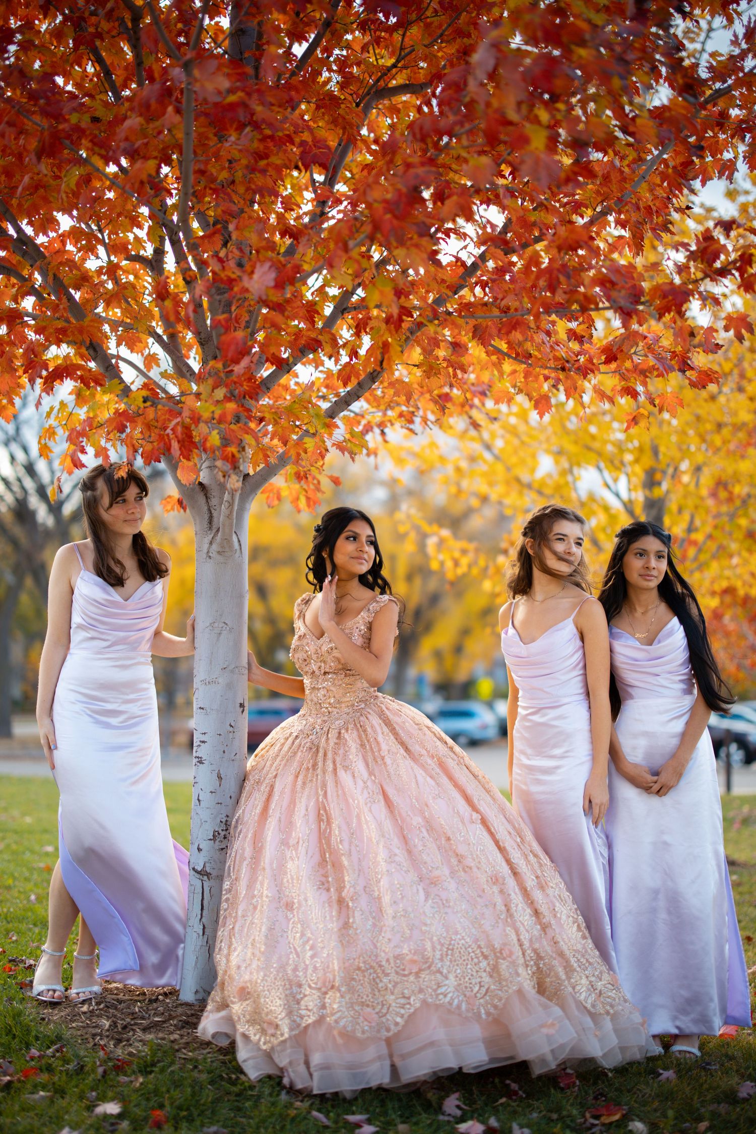 Quinceañera girl in rose gold ball gown stands with three ladies in white dresses under vibrant red autumn tree.
