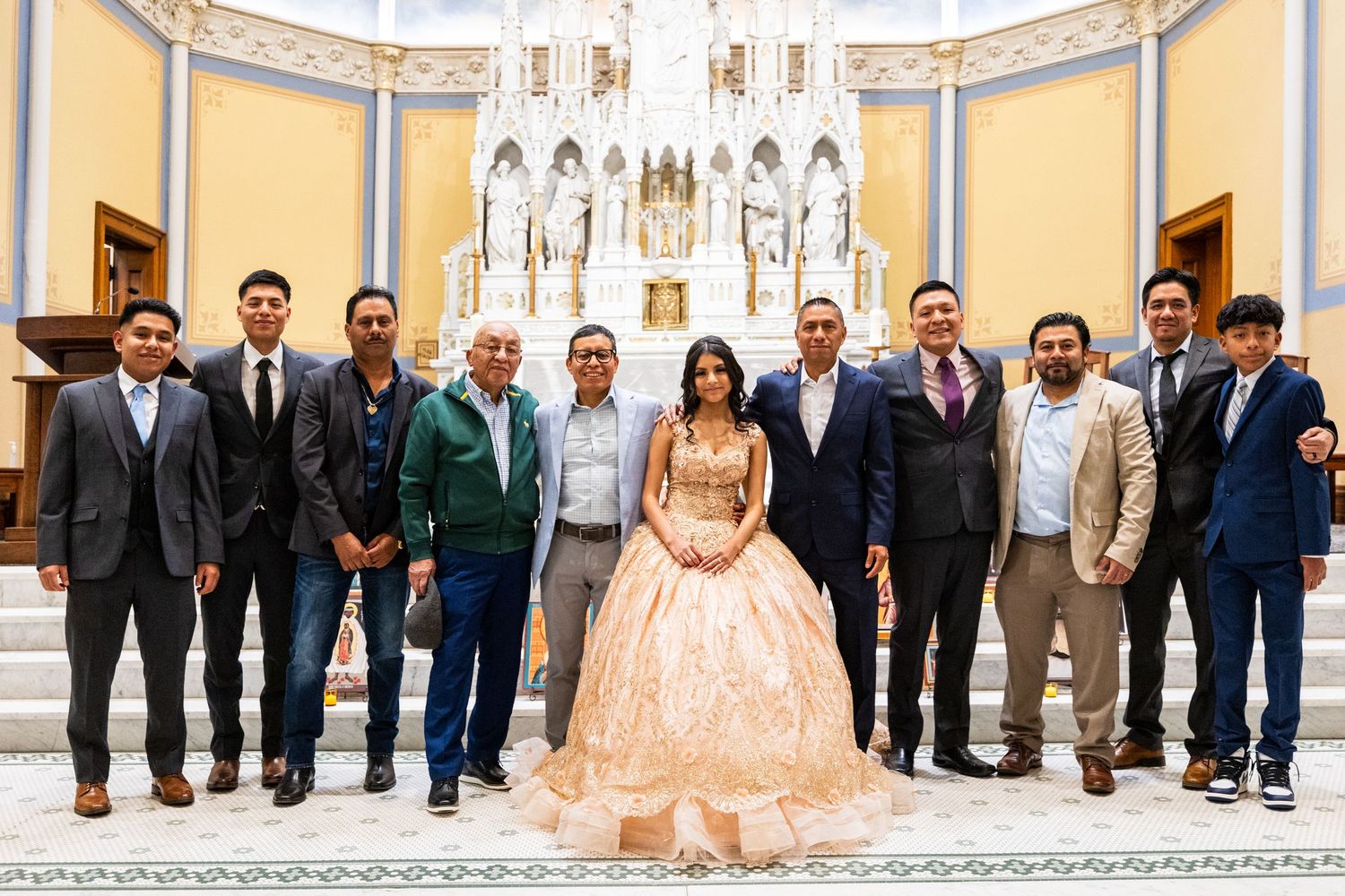 Quinceañera celebrant in peach gown poses with group of men in suits inside decorated Catholic church.