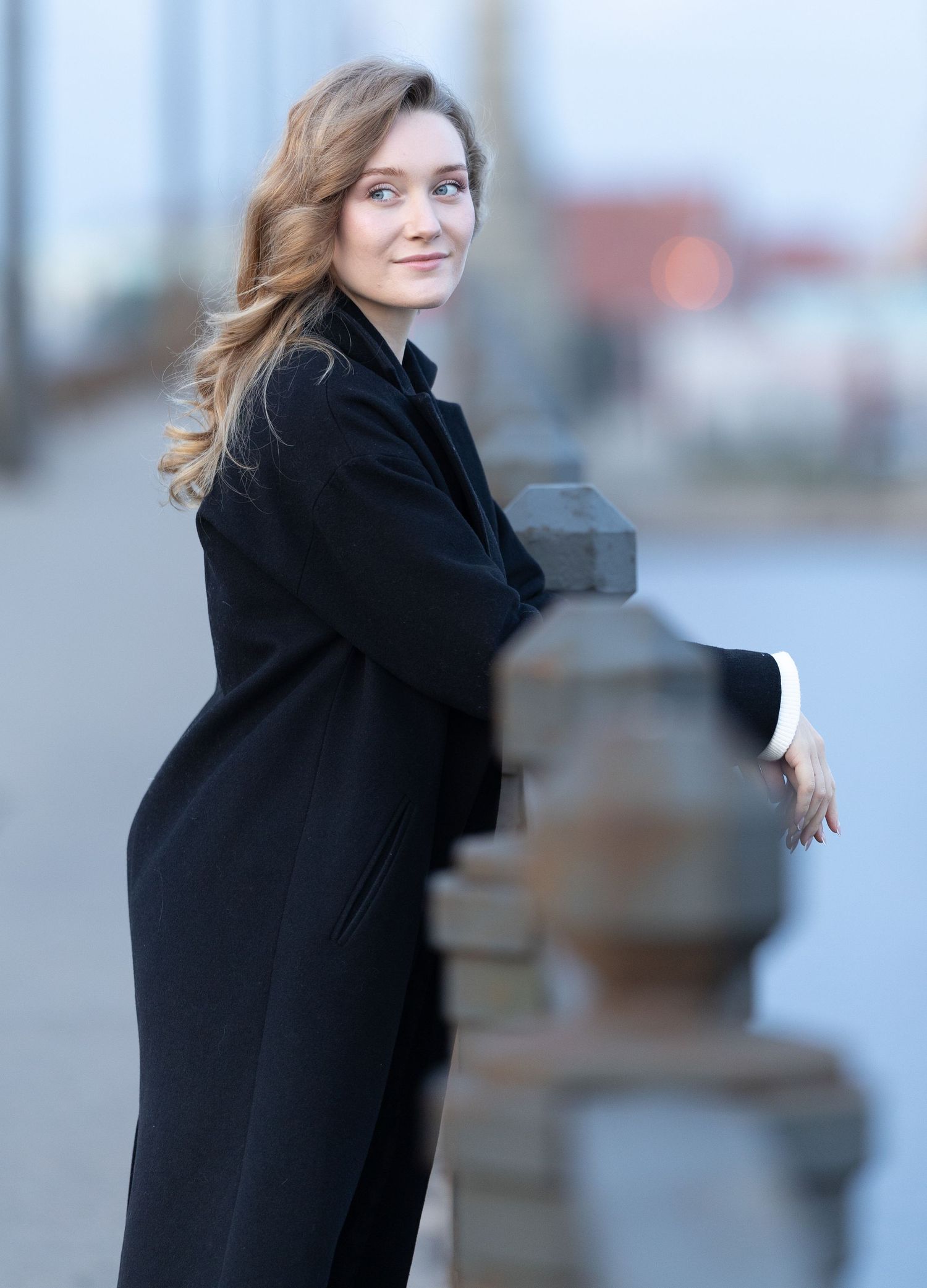 Elegant woman in black coat leaning on stone railing, smiling over shoulder outdoors.