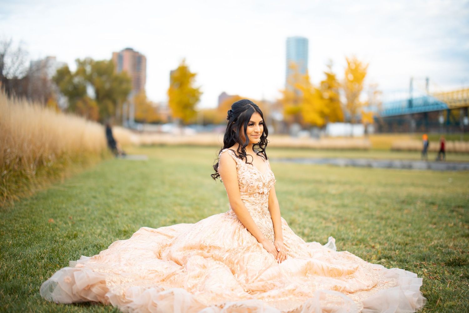 Beautiful quinceañera girl in ornate rose gold ball gown sits gracefully on green grass with city skyline behind.