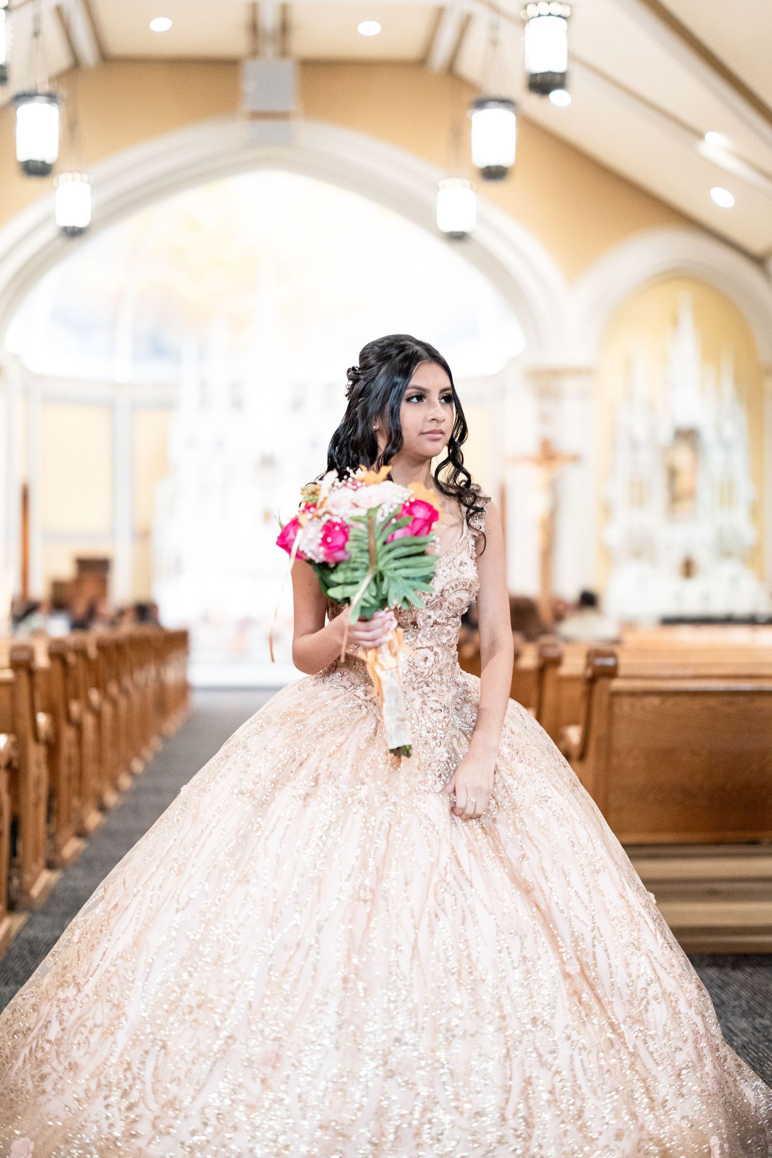 Young woman in ornate peach quinceañera ball gown holds floral bouquet inside beautiful church aisle.