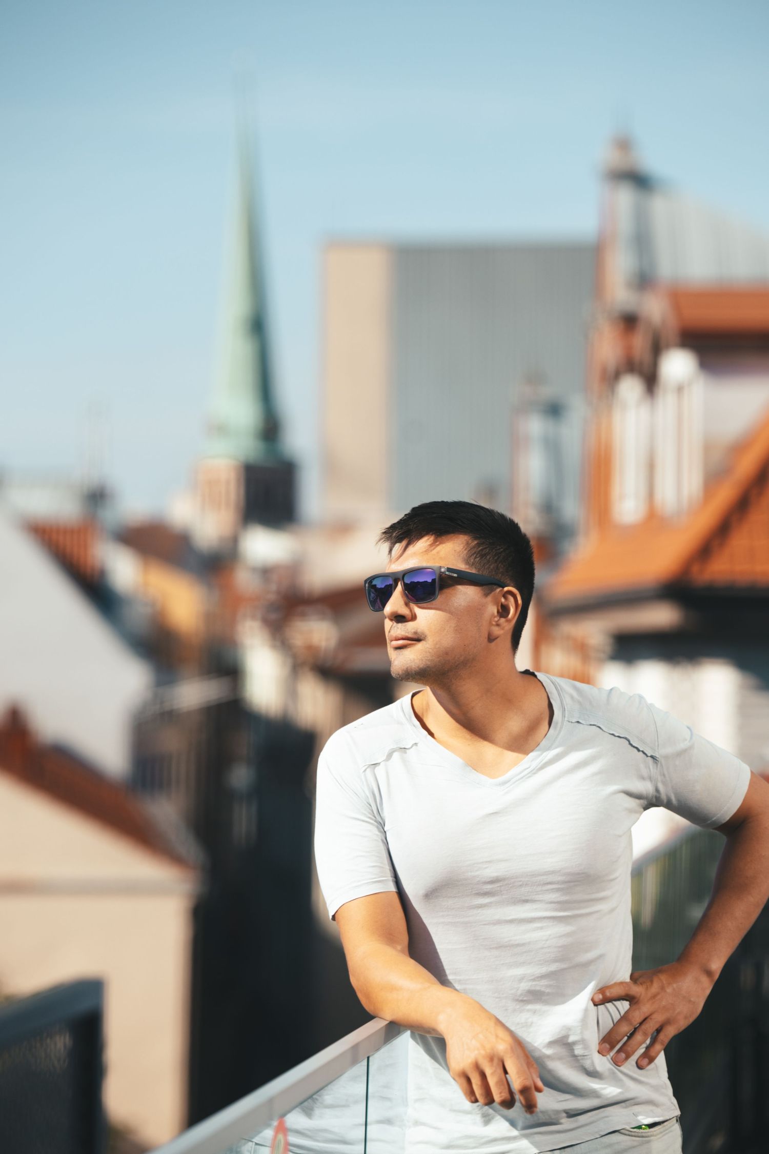Man in white t-shirt and sunglasses leaning on a railing with European city rooftops in the background.