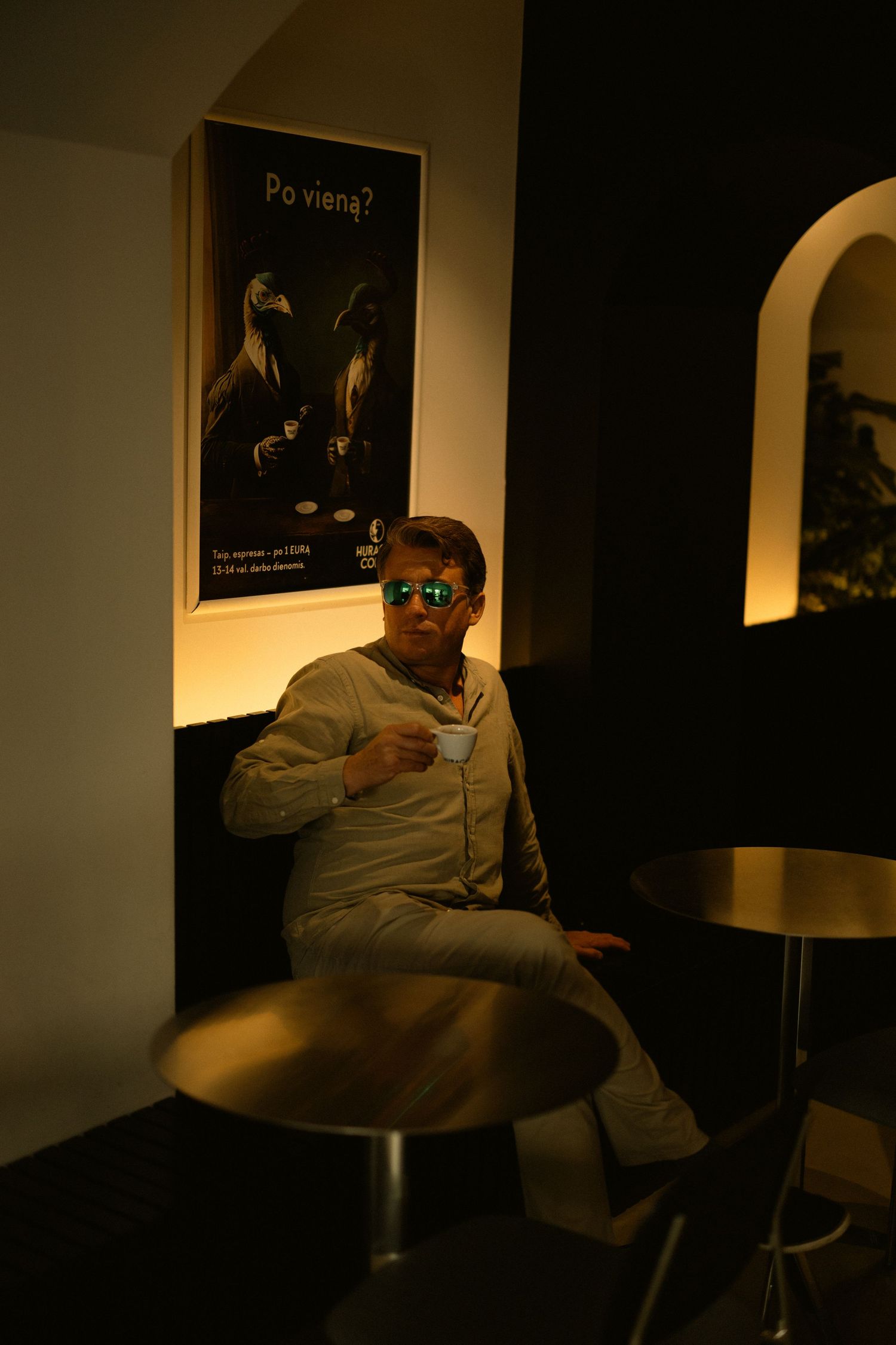Man in beige outfit wearing mirrored sunglasses sitting in dark cafe holding coffee cup near poster.