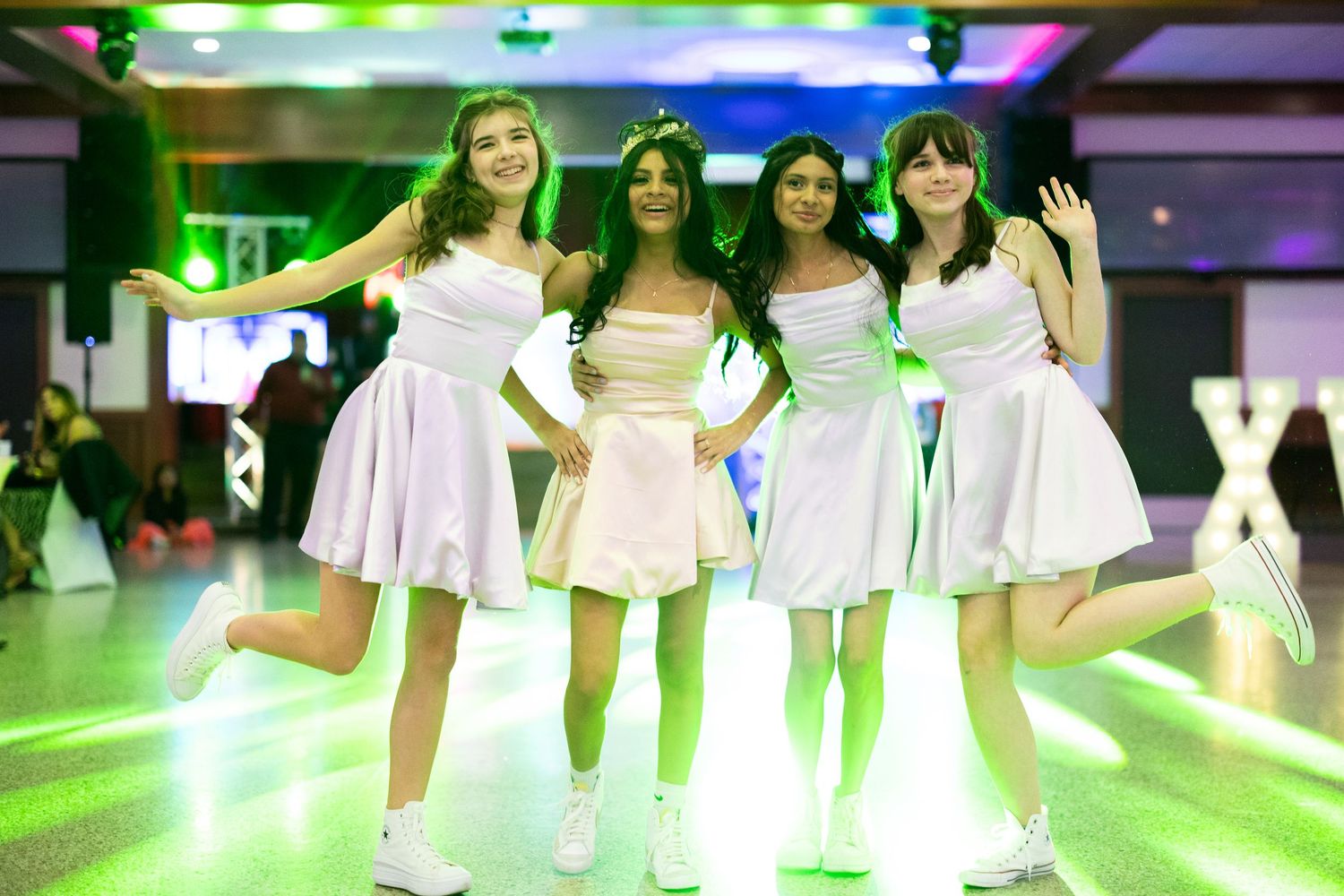 Four girls in matching white dresses pose together under green lights at a quinceañera celebration.